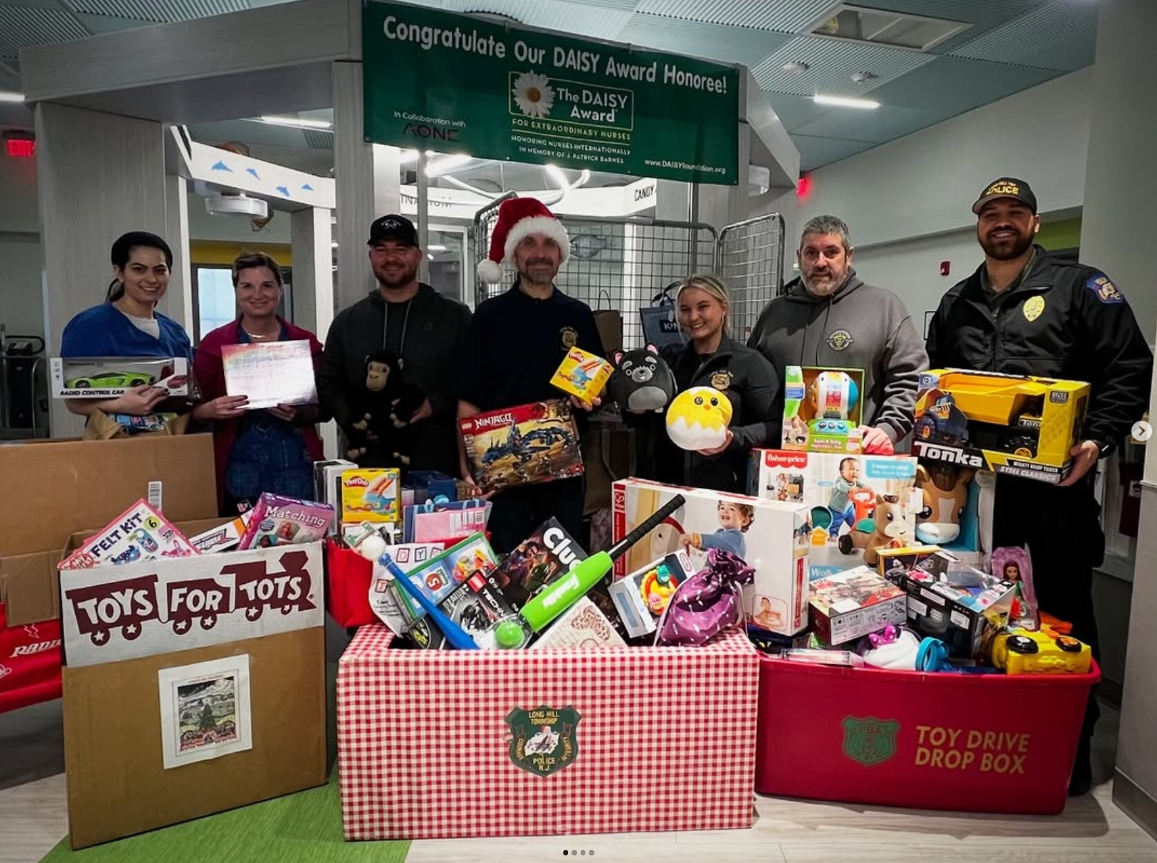 Group of six people, including two police officers, standing behind boxes filled with toys for a holiday toy drive in a community center.
