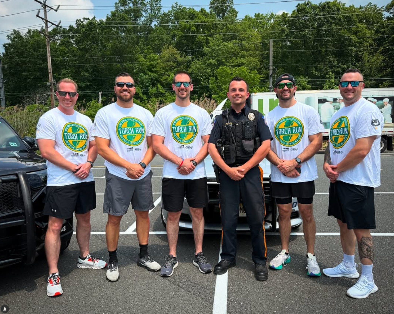 Group of five men in white Torch Run t-shirts and one police officer posing together in a parking lot.