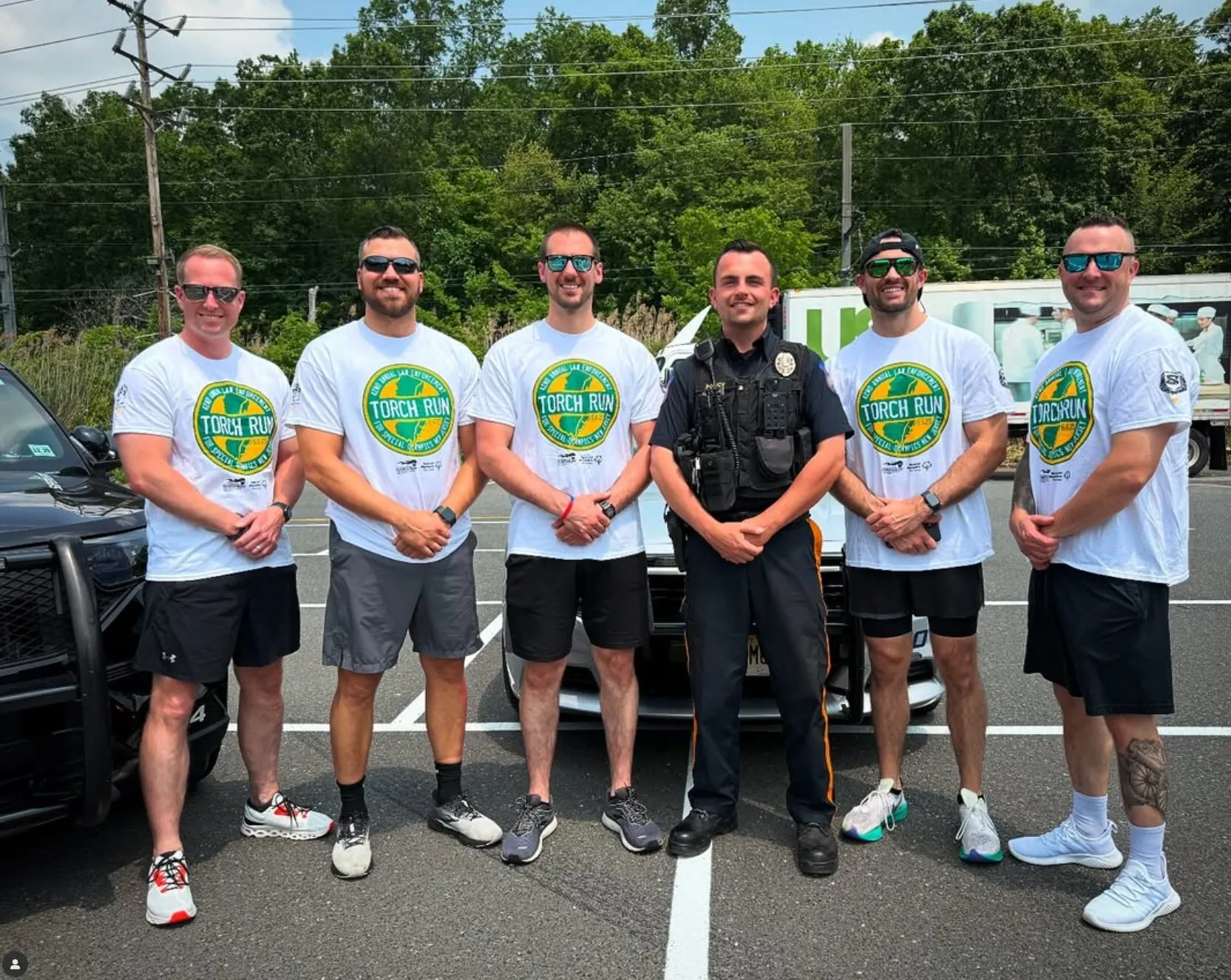 Group of five men in white Torch Run t-shirts and one police officer posing together in a parking lot.