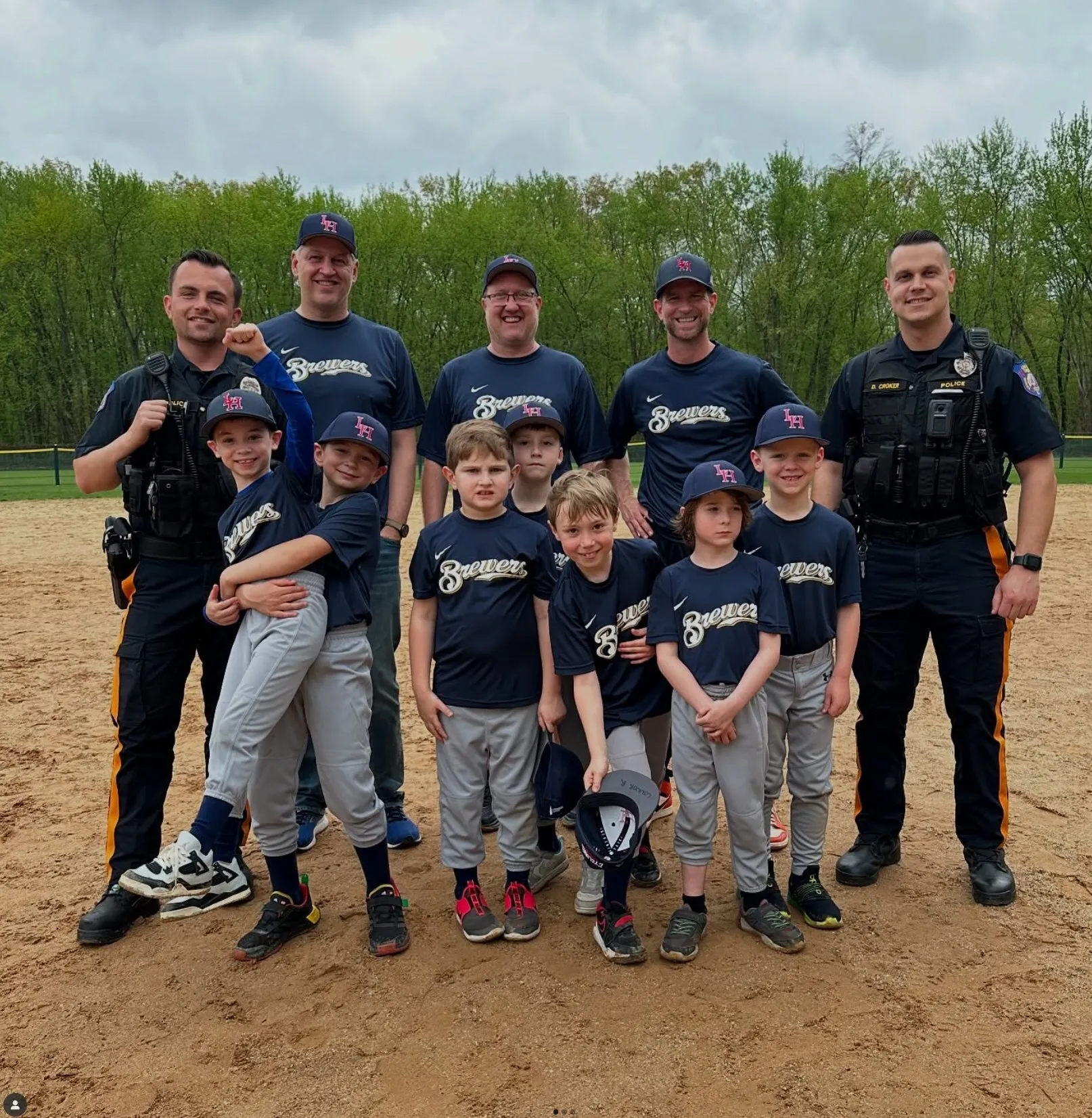 Youth baseball team in navy blue 'Brewers' jerseys posing with four police officers on a baseball field.
