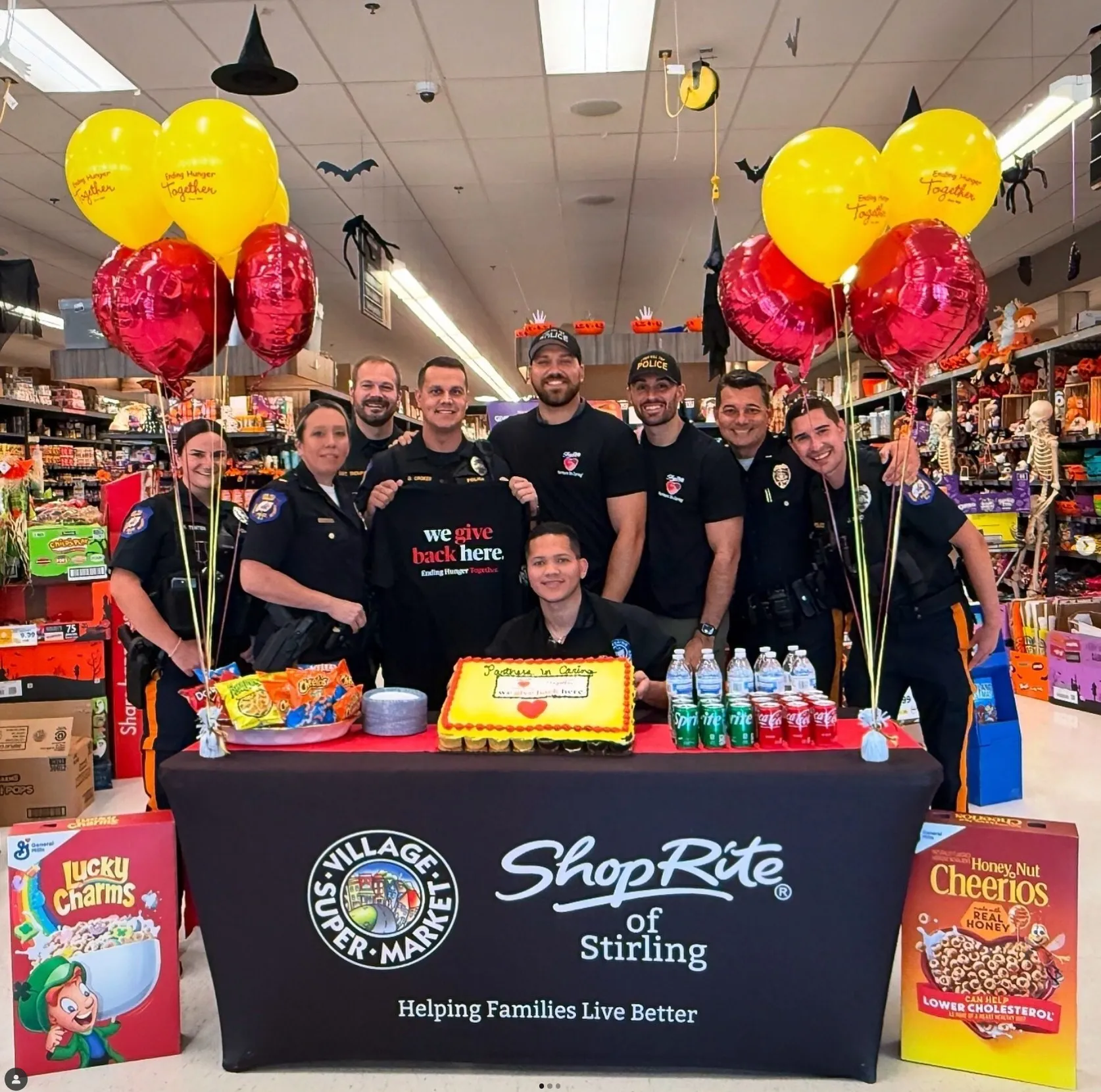 Group of police officers and volunteers posing behind a ShopRite of Stirling table with a cake, drinks, snacks, and balloons in a supermarket decorated for Halloween.