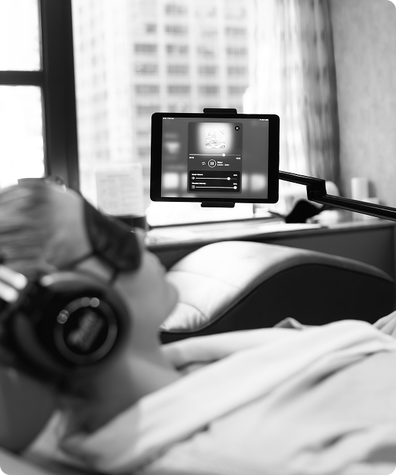 Person reclining on a chair wearing headphones, watching a tablet mounted on a flexible arm in a room with large windows.