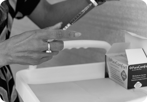 Hand holding a syringe with a measurement scale near a white table that has a box of PureComfort alcohol prep pads.