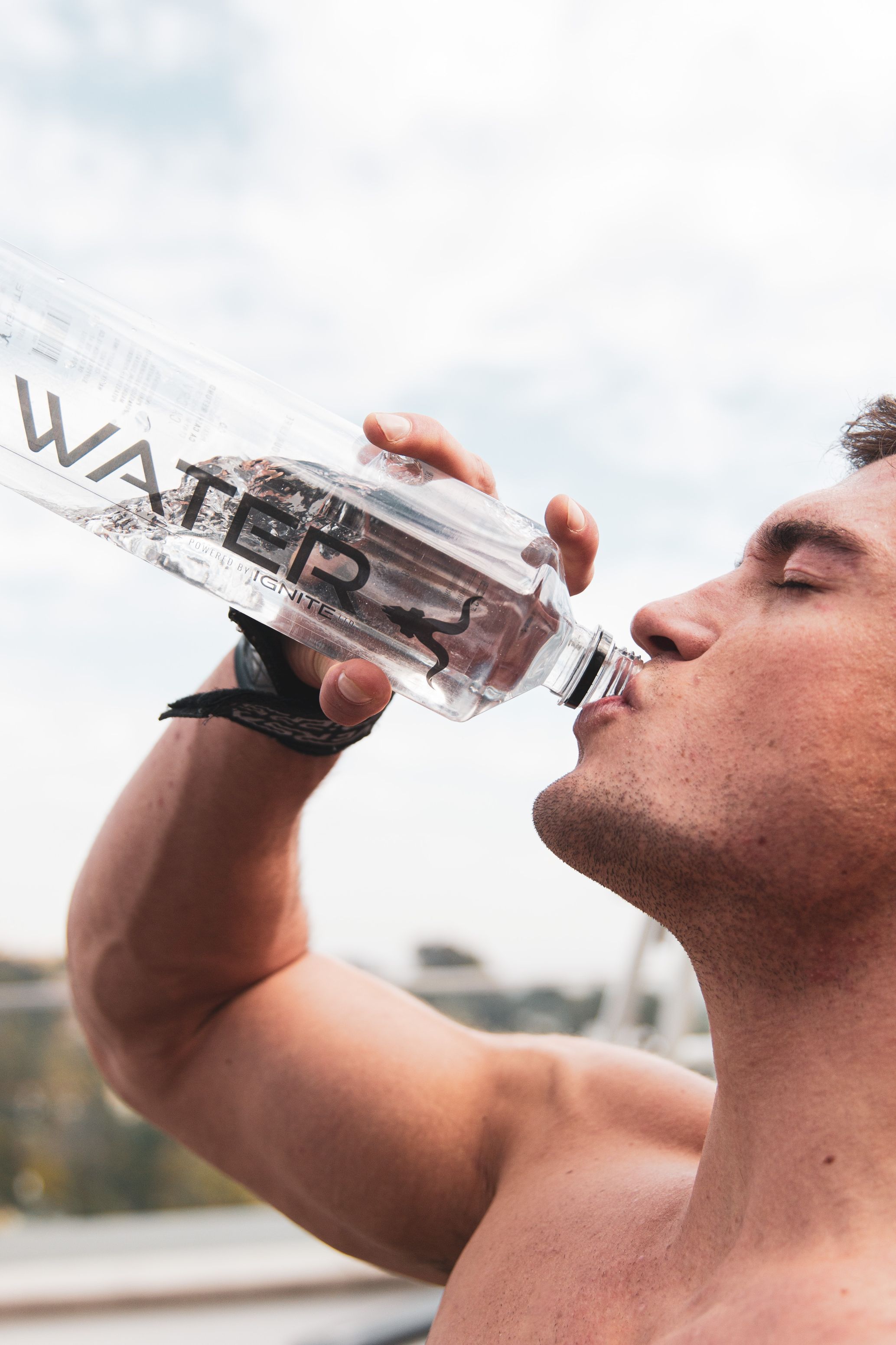 Muscular shirtless man drinking water from a clear bottle labeled 'WATER' outdoors with a cloudy sky background.