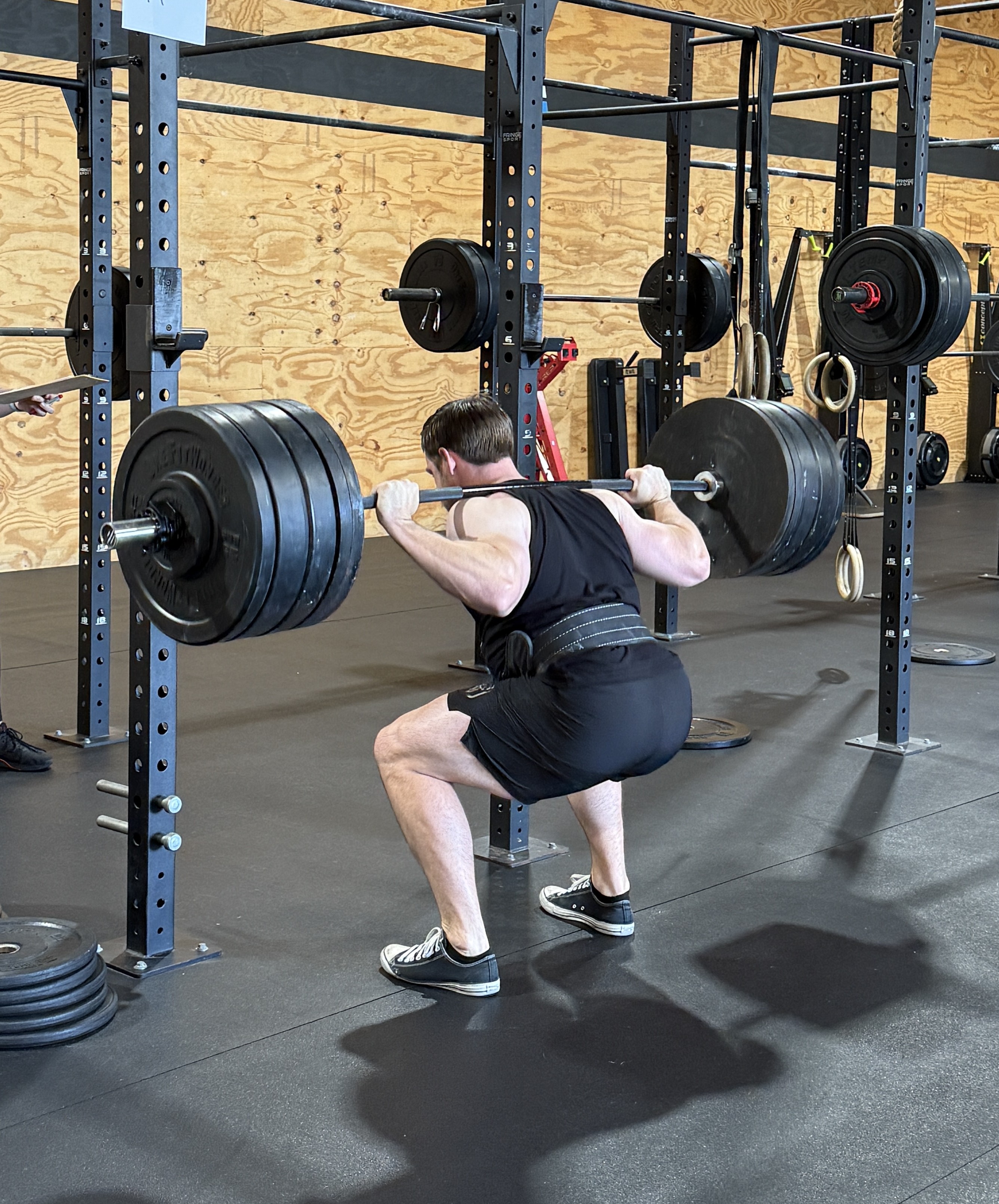 Man wearing a black tank top and shorts performing a heavy barbell back squat in a gym with wooden walls and black rubber flooring.