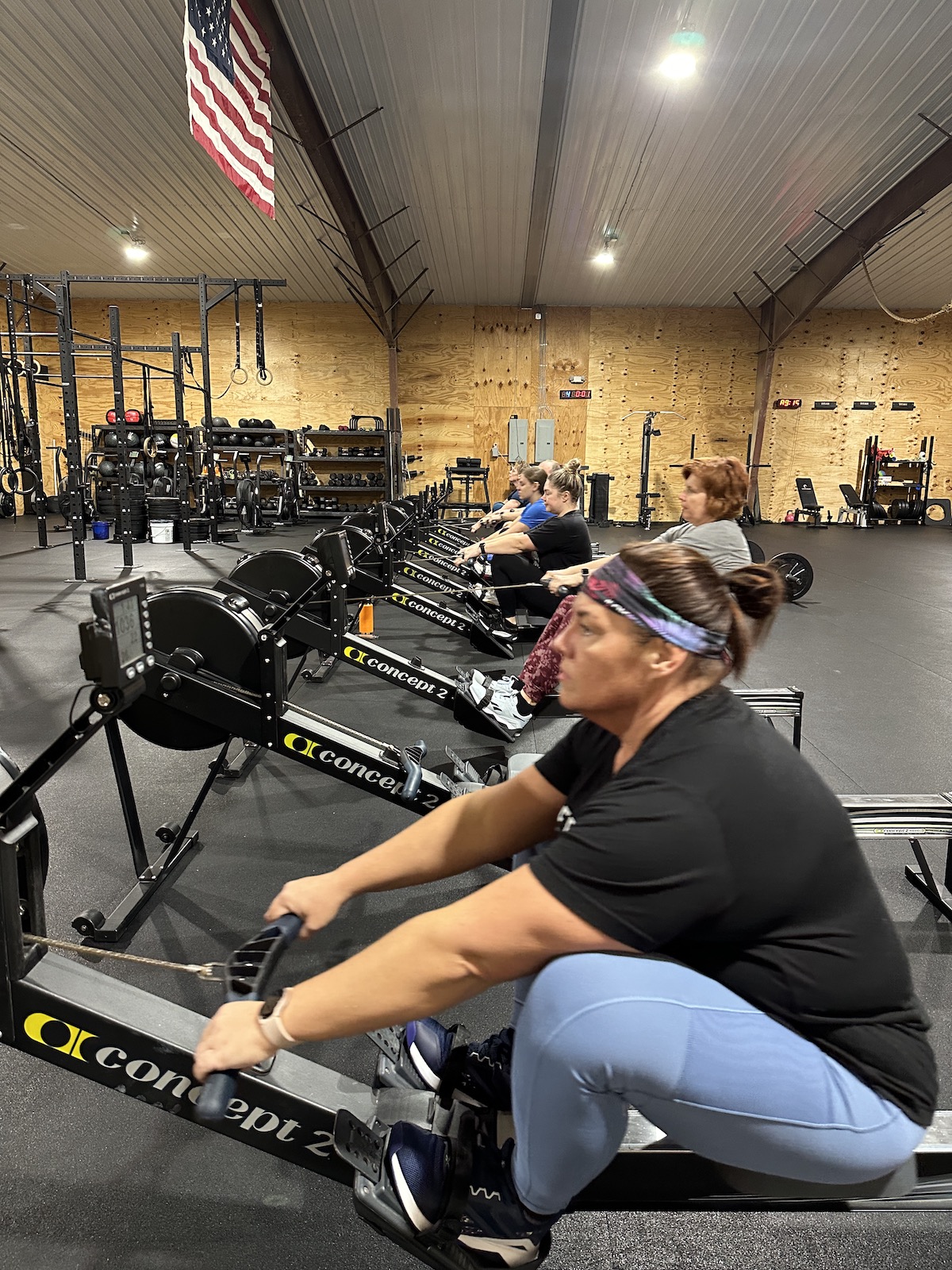 Group of women exercising on Concept2 rowing machines in a gym with an American flag hanging from the ceiling.