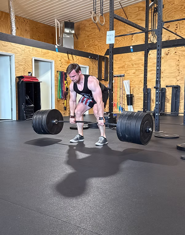 Man wearing tank top and shorts lifting a heavily weighted barbell in a gym with wooden walls and fitness equipment.