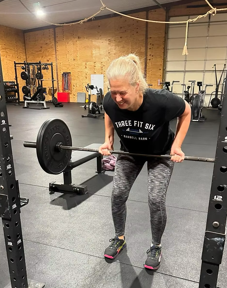 Smiling woman lifting a barbell in a gym with wooden walls and exercise equipment in the background.