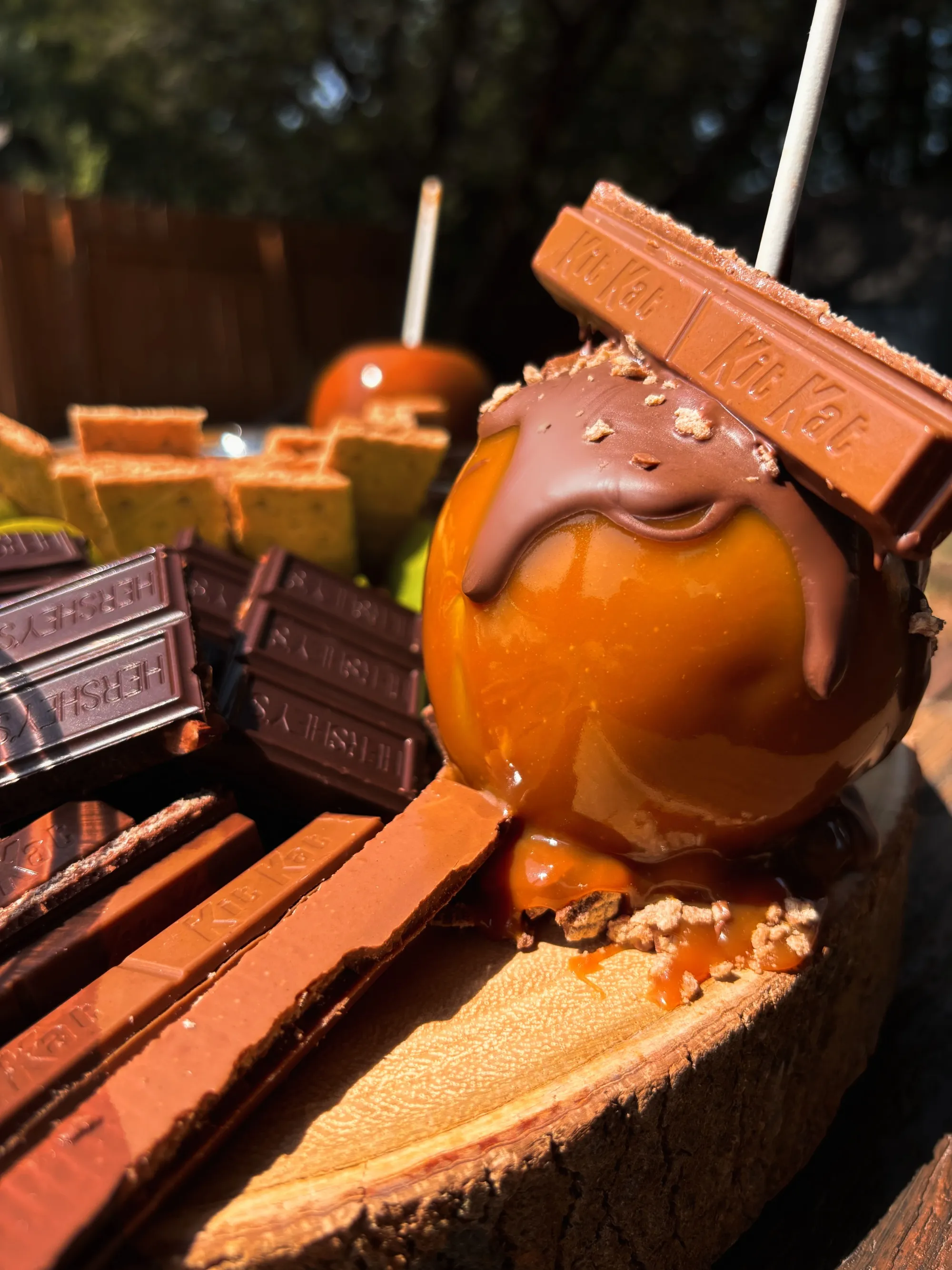 Close-up of caramel apples topped with melted chocolate and Kit Kat bars, with Hershey’s chocolate pieces and graham crackers on a wooden board.