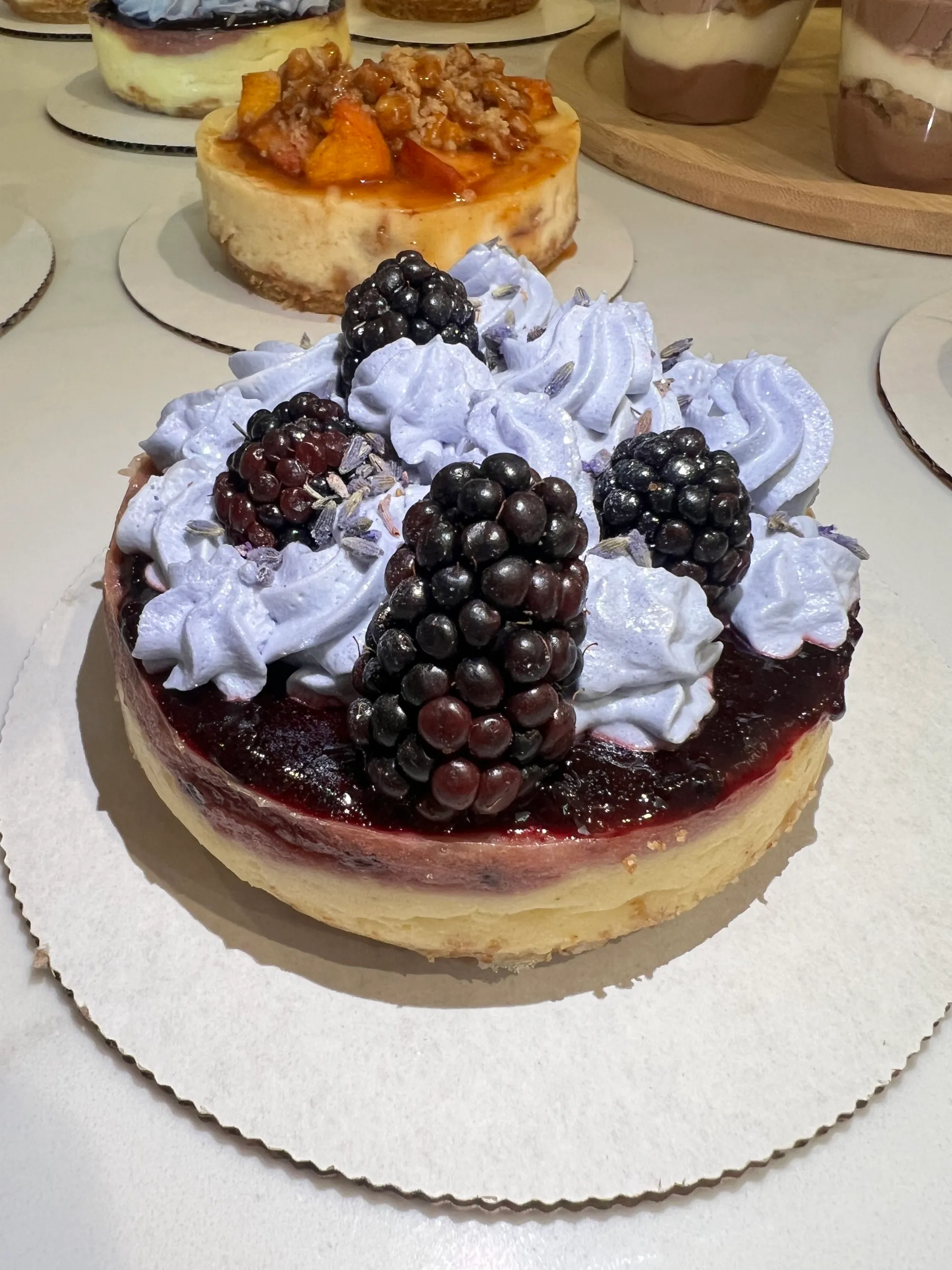 Cheesecake topped with blackberries, purple whipped cream, and lavender buds on a white cardboard base.