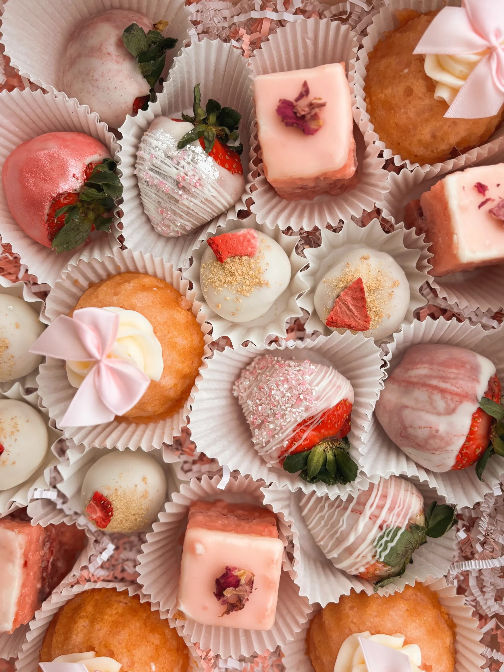 Assorted desserts including white chocolate-covered strawberries with pink sprinkles, cupcakes topped with pink bows, and pink frosted petit fours placed in paper cups.