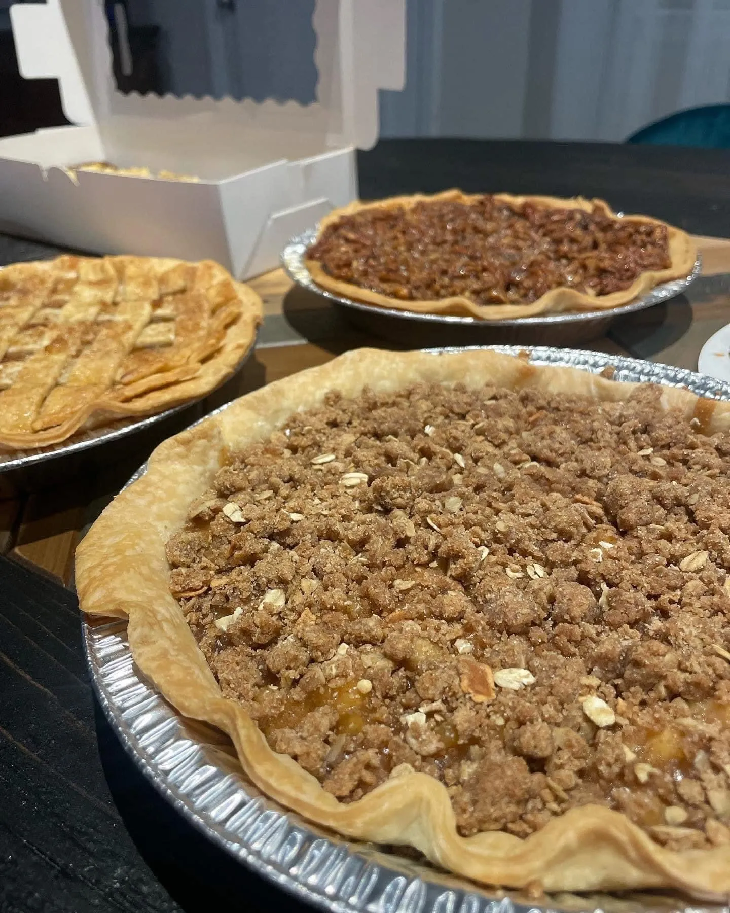 Three homemade pies on a table, including an oatmeal crumble pie in the foreground, a pecan pie in the back, and a lattice-topped pie on the left.