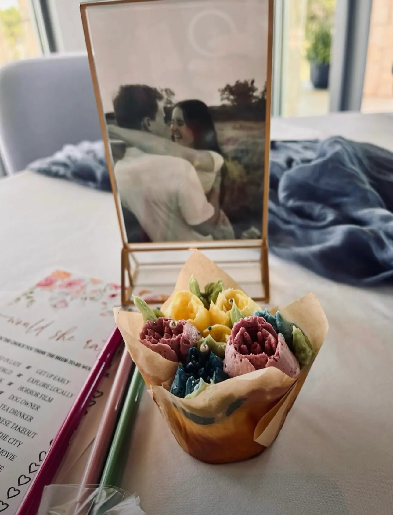 A bouquet of colorful crepe paper flowers sits on a table with colored pencils and a framed black-and-white photo of a couple embracing in the background.