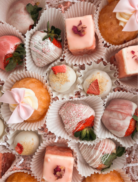 Assorted elegant desserts including pink frosted cakes, white chocolate-covered strawberries, and cupcakes with pink bows in white paper cups.