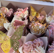 Assorted decorated cupcakes with pink frosting, gold and pink toppers including a butterfly, chocolate pieces, and a crown.