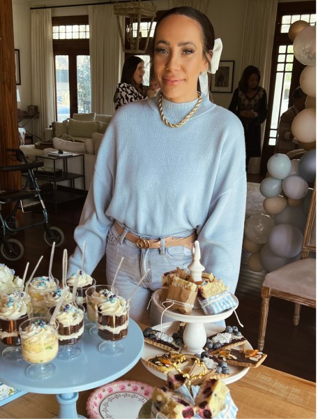 Woman in light blue sweater and gold necklace smiling behind a table with assorted desserts and cupcakes.