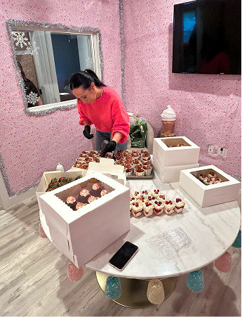 Woman in a pink sweater arranging various cupcakes and desserts on a round table with pink glittery walls in the background.