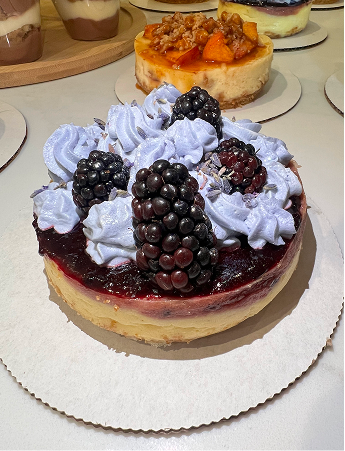 Cheesecake topped with blackberries and purple whipped cream on a white paper base.