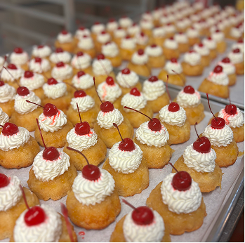 Rows of small bundt cakes topped with whipped cream and a single cherry on each cake on a tray.