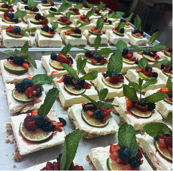 Squares of layered cheesecake topped with fresh mixed berries, lime slices, and mint leaves arranged on trays.