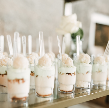 Multiple clear plastic cups filled with layered creamy dessert topped with a scoop and a clear spoon on a reflective surface.