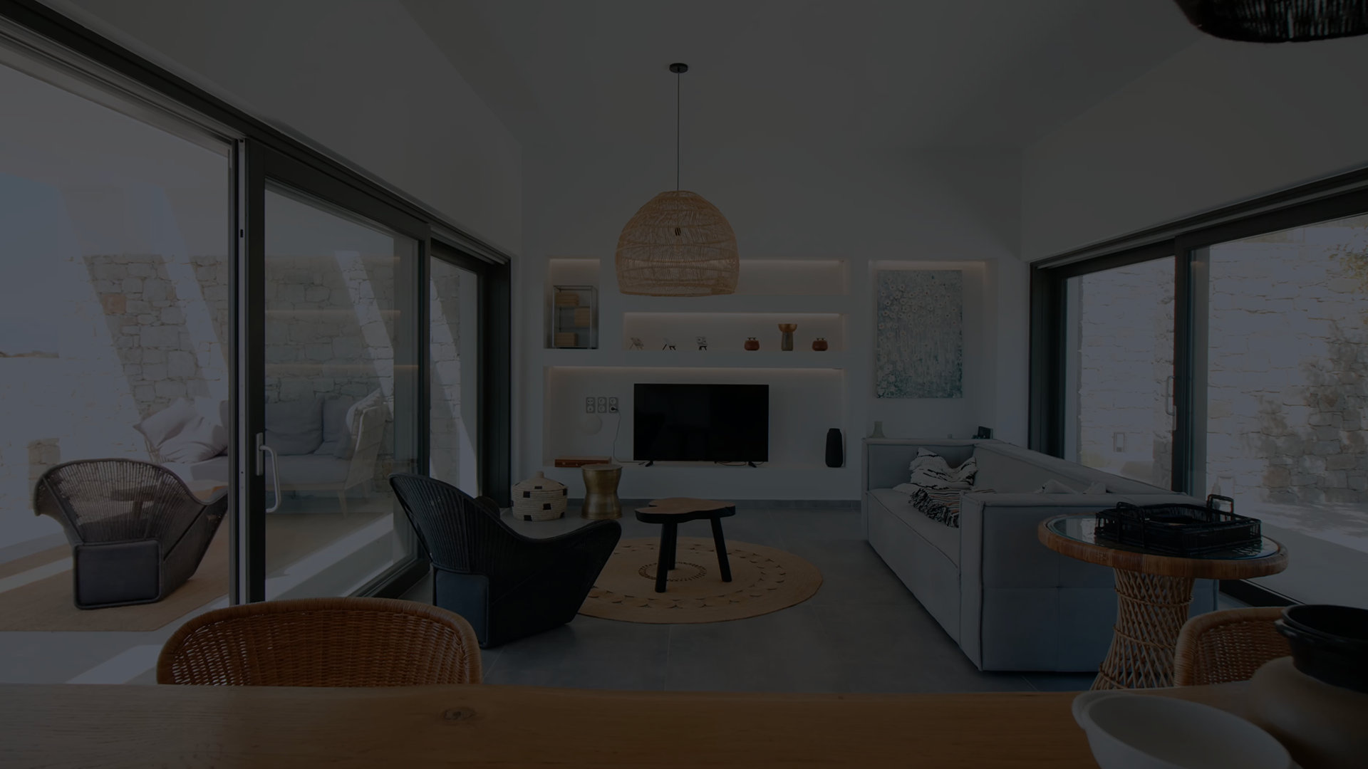 Salon moderne avec grandes baies vitrées, canapé gris, fauteuils en osier noirs, table basse ronde et étagères murales éclairées avec télévision.