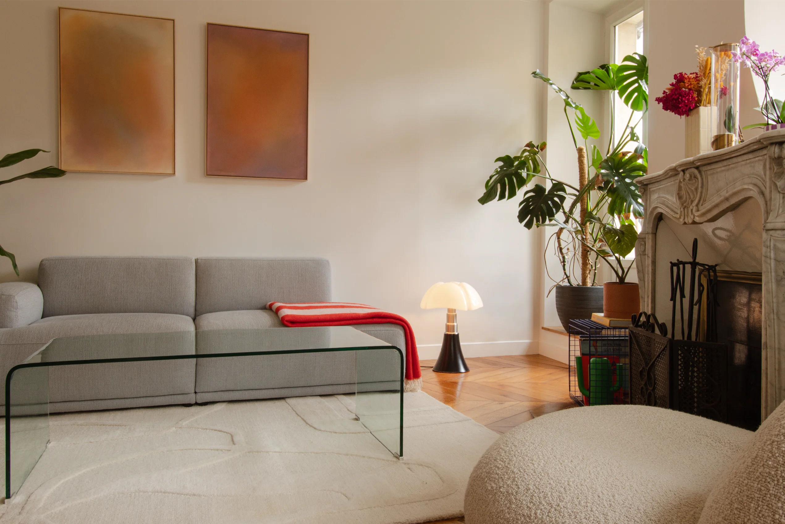 Salon minimaliste avec un canapé gris, une table basse en verre, un tapis blanc texturé, une lampe moderne, des plantes en pot et une cheminée en pierre.
