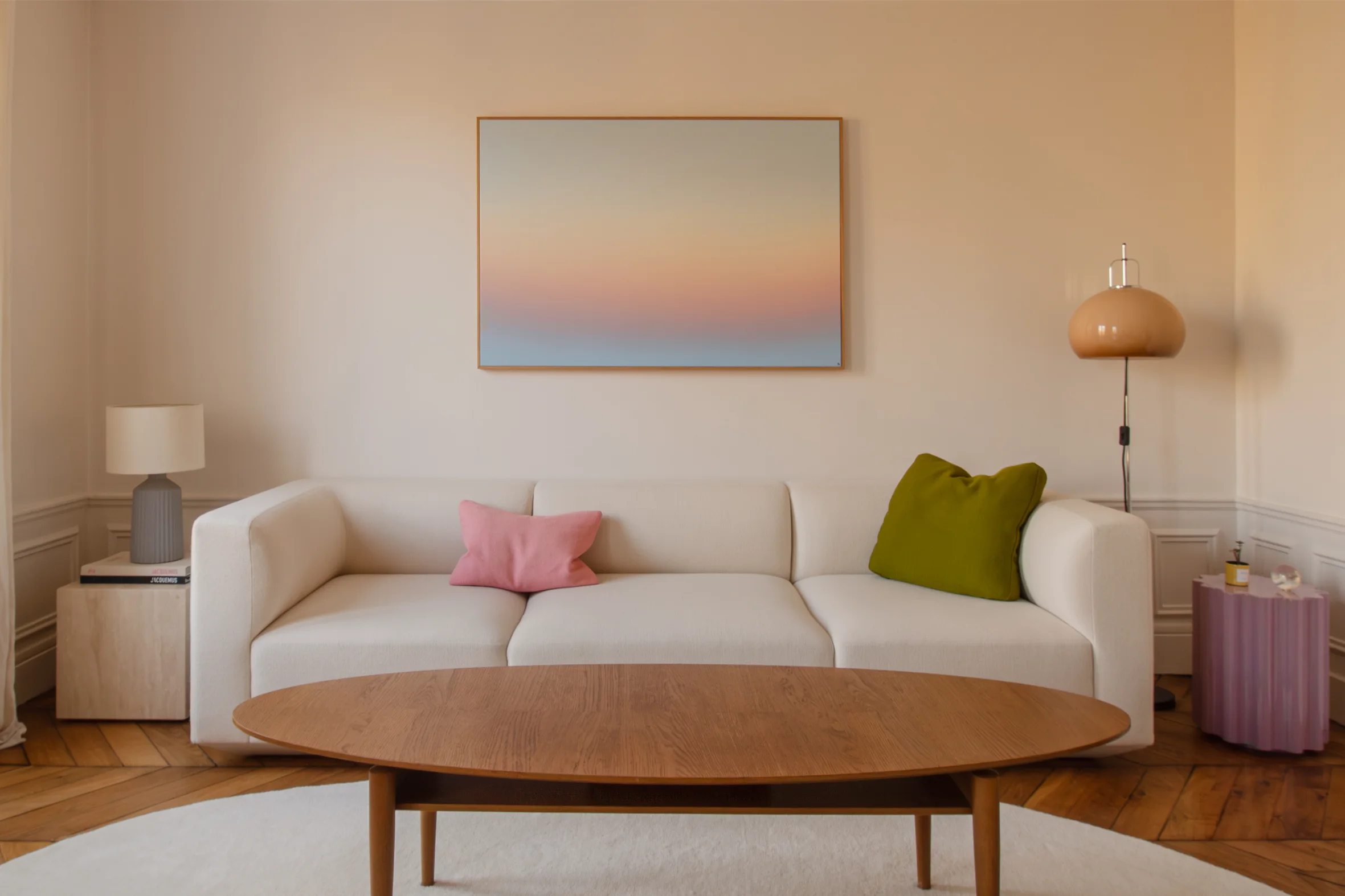 Salon minimaliste avec un canapé crème, des coussins roses et verts, une table basse en bois, des tables d'appoint avec des lampes et une œuvre d'art représentant un coucher de soleil dégradé.