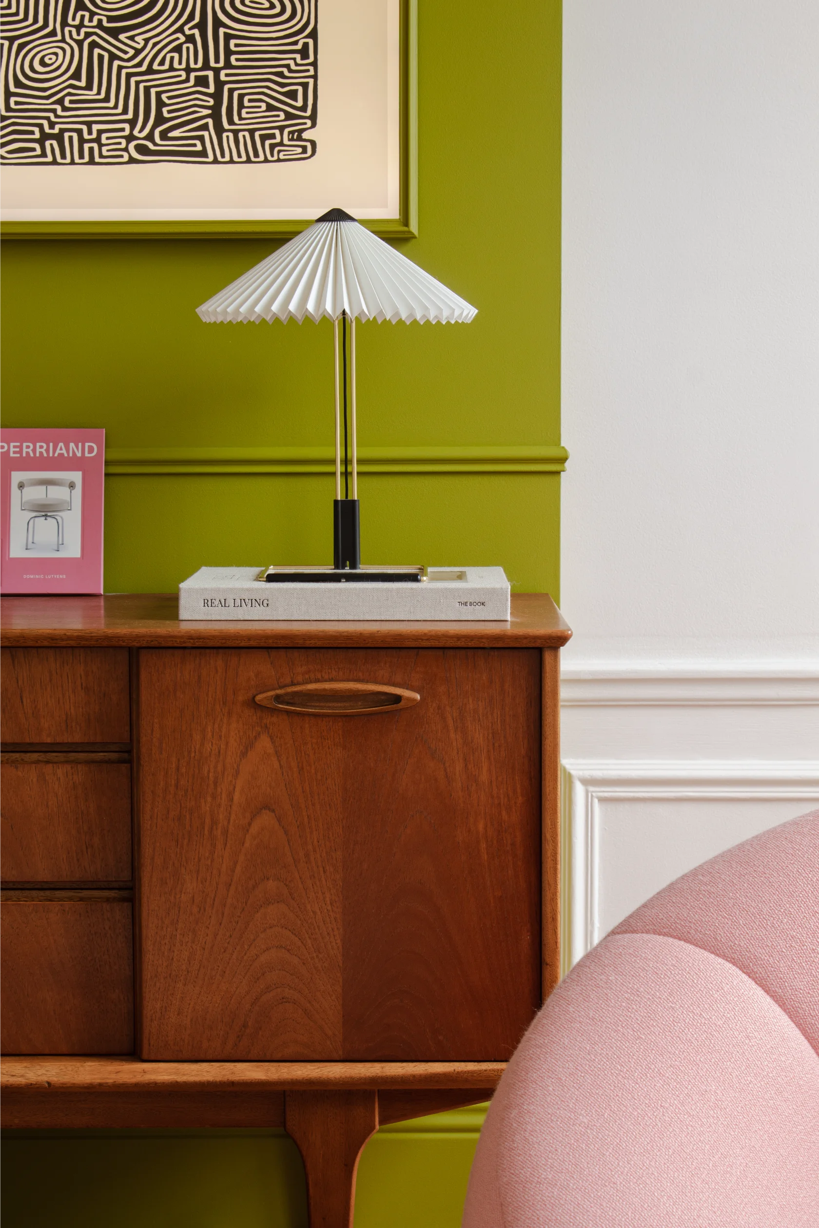 Buffet en bois du milieu du siècle avec une lampe de table blanche plissée, un livre rose intitulé Perriand et une œuvre d'art abstraite en noir et blanc encadrée sur un mur d'accent vert, à côté d'une chaise courbée rose.