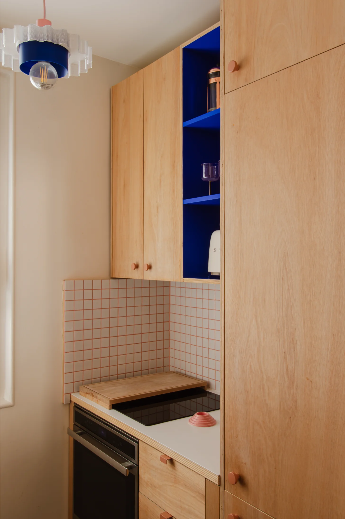 Coin cuisine moderne avec des armoires en bois clair, des étagères ouvertes bleues, un dosseret carrelé, une plaque de cuisson noire et une planche à découper.