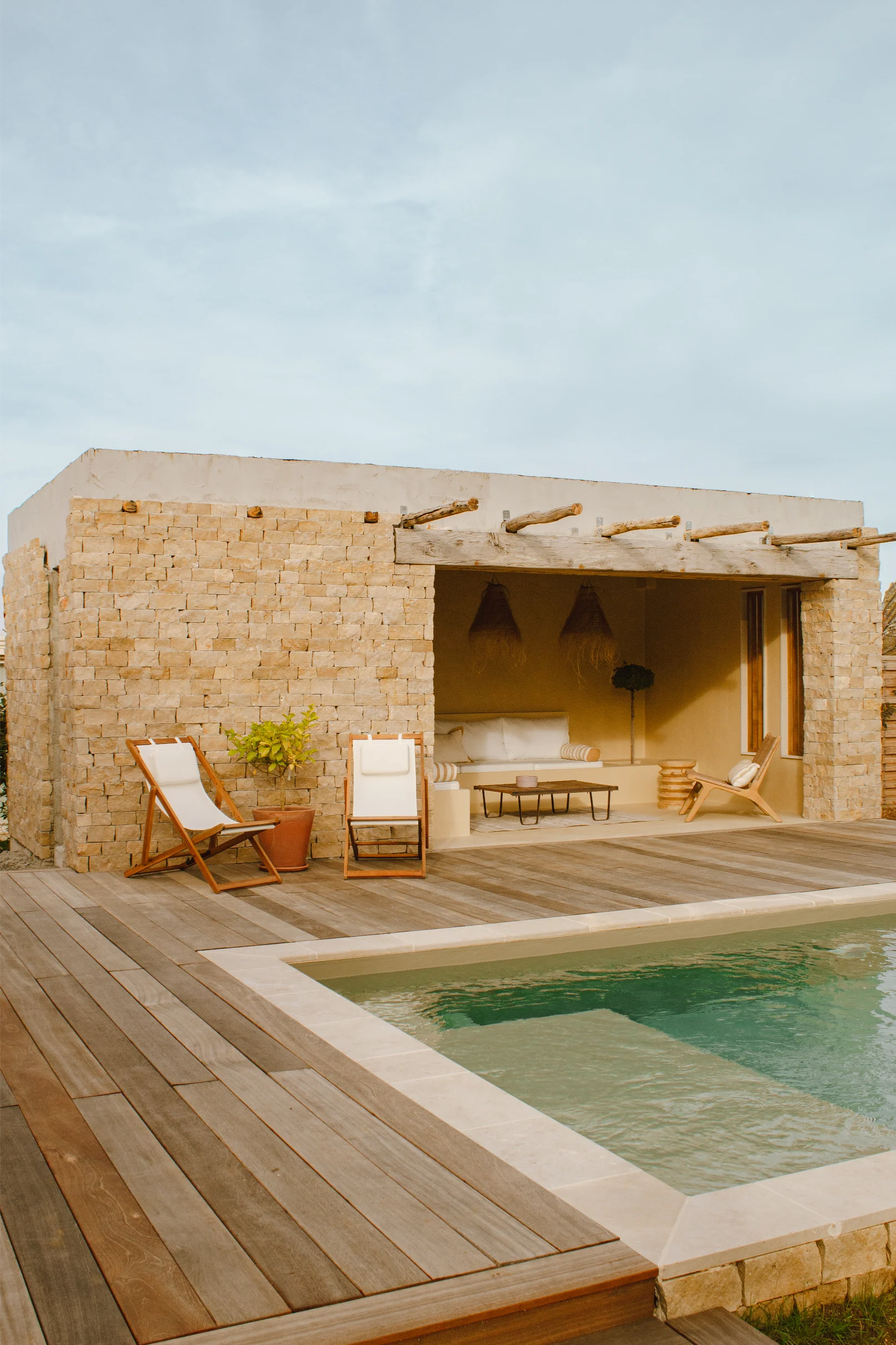 Patio extérieur moderne avec terrasse en bois, deux chaises longues en toile blanche, une plante en pot et une piscine devant un bâtiment en pierre avec un coin salon ombragé.