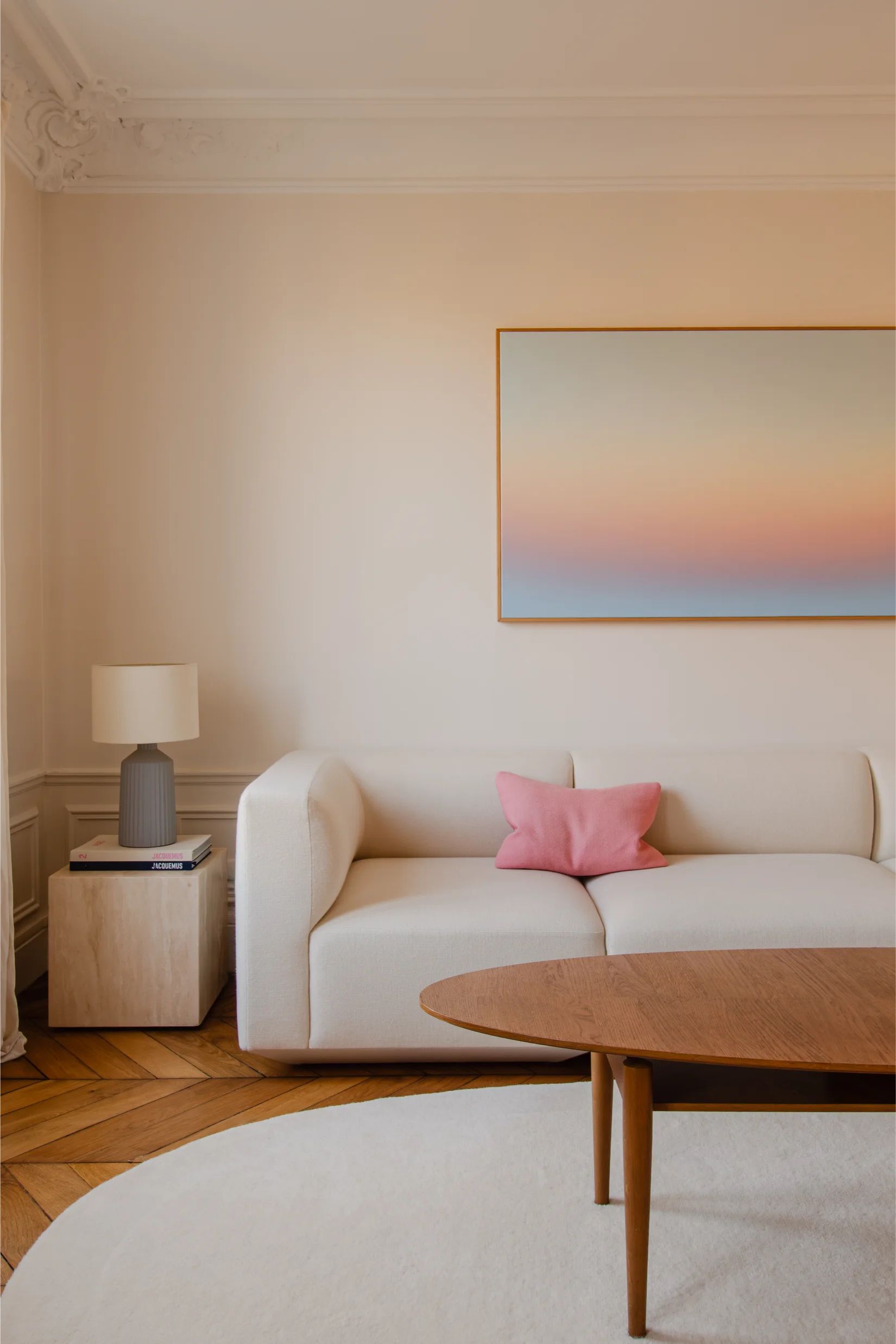 Salon minimaliste avec un canapé blanc, un coussin rose, une table basse en bois, une table d'appoint avec une lampe et une œuvre d'art représentant un coucher de soleil dégradé.