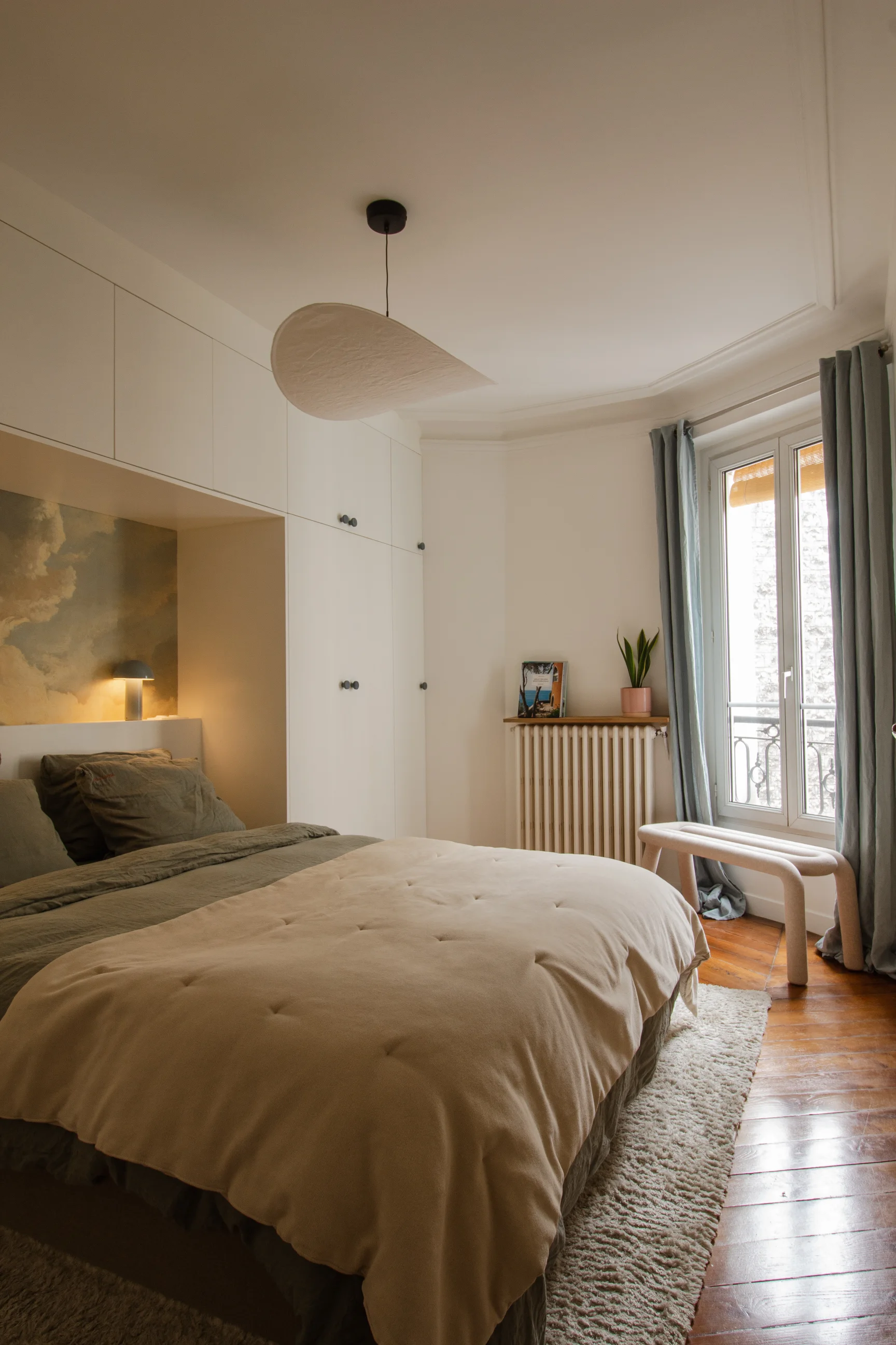 Chambre confortable avec literie beige et olive, armoires encastrées blanches, fresque murale représentant des nuages, parquet et fenêtre avec rideaux bleus.