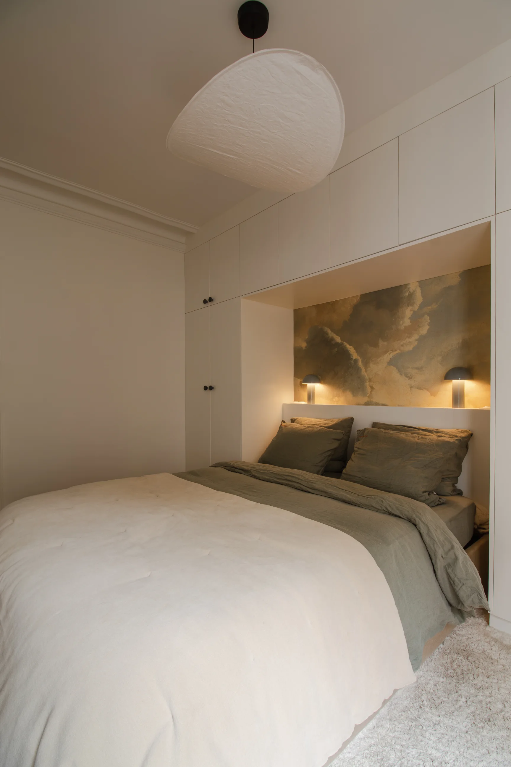 Chambre confortable avec des armoires encastrées blanches entourant le lit, une literie verte, une tête de lit sur le thème des nuages et un éclairage tamisé.