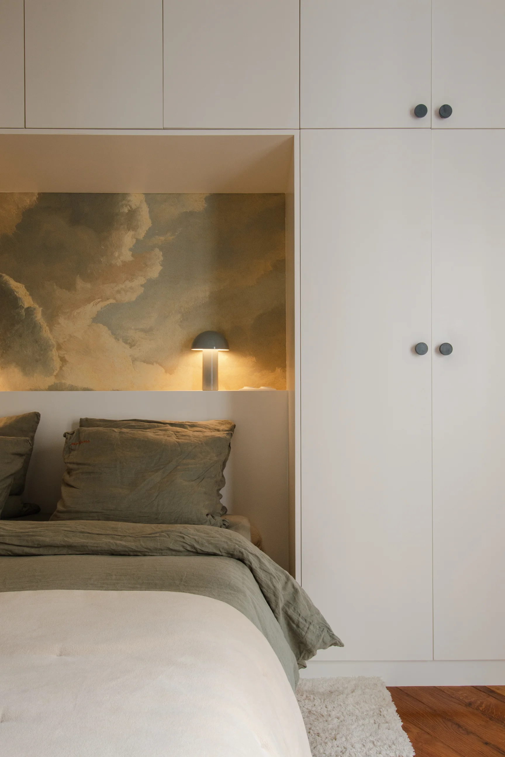 Coin chambre avec armoire encastrée blanche, lit avec literie verte et petite lampe de table éclairant une niche murale décorée d'un motif nuageux.