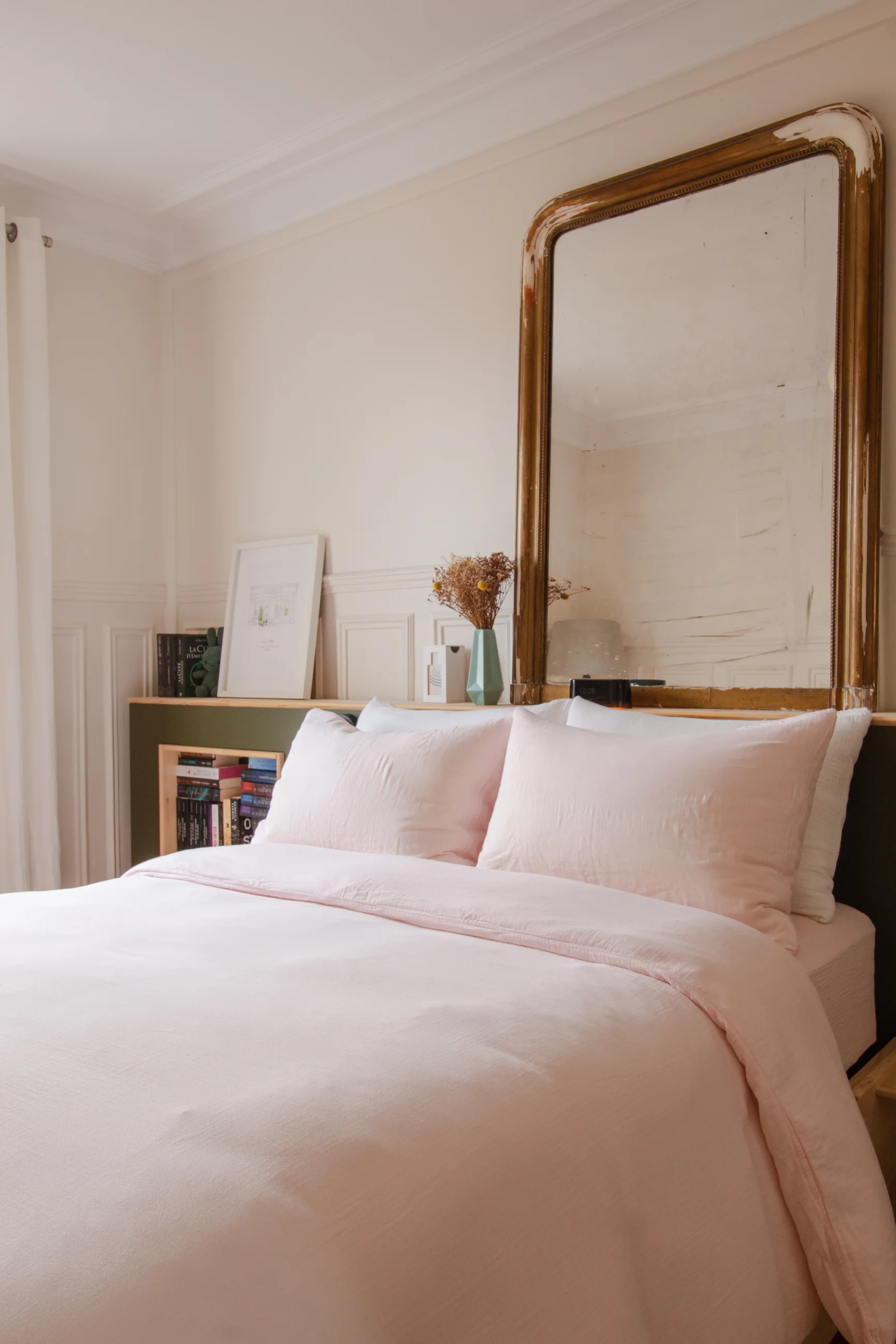 Chambre à coucher avec literie rose pâle, miroir antique à cadre doré et bibliothèque décorative.