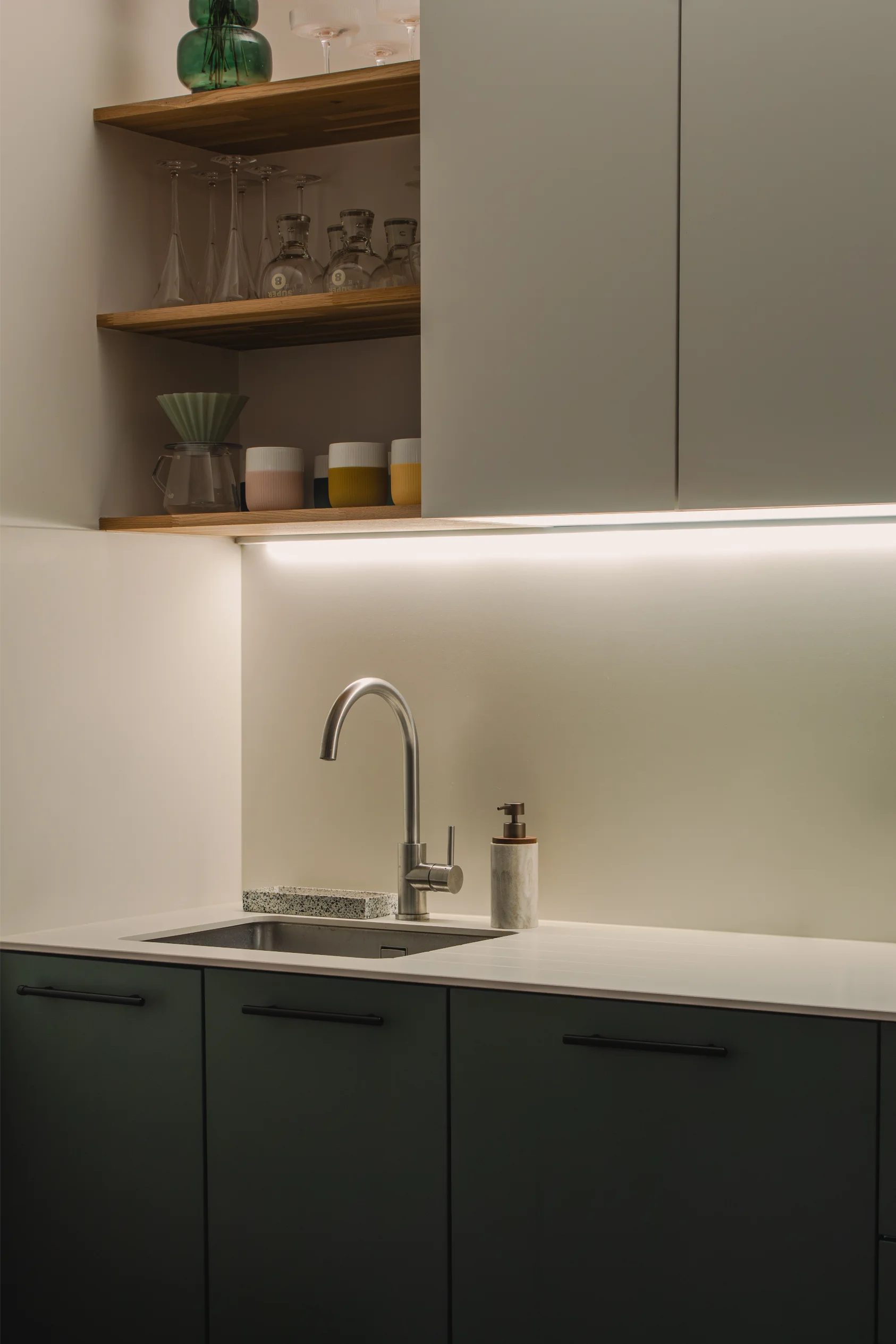 Évier de cuisine moderne avec robinet en acier inoxydable, éclairage sous les armoires et étagères pour ranger verres et tasses.