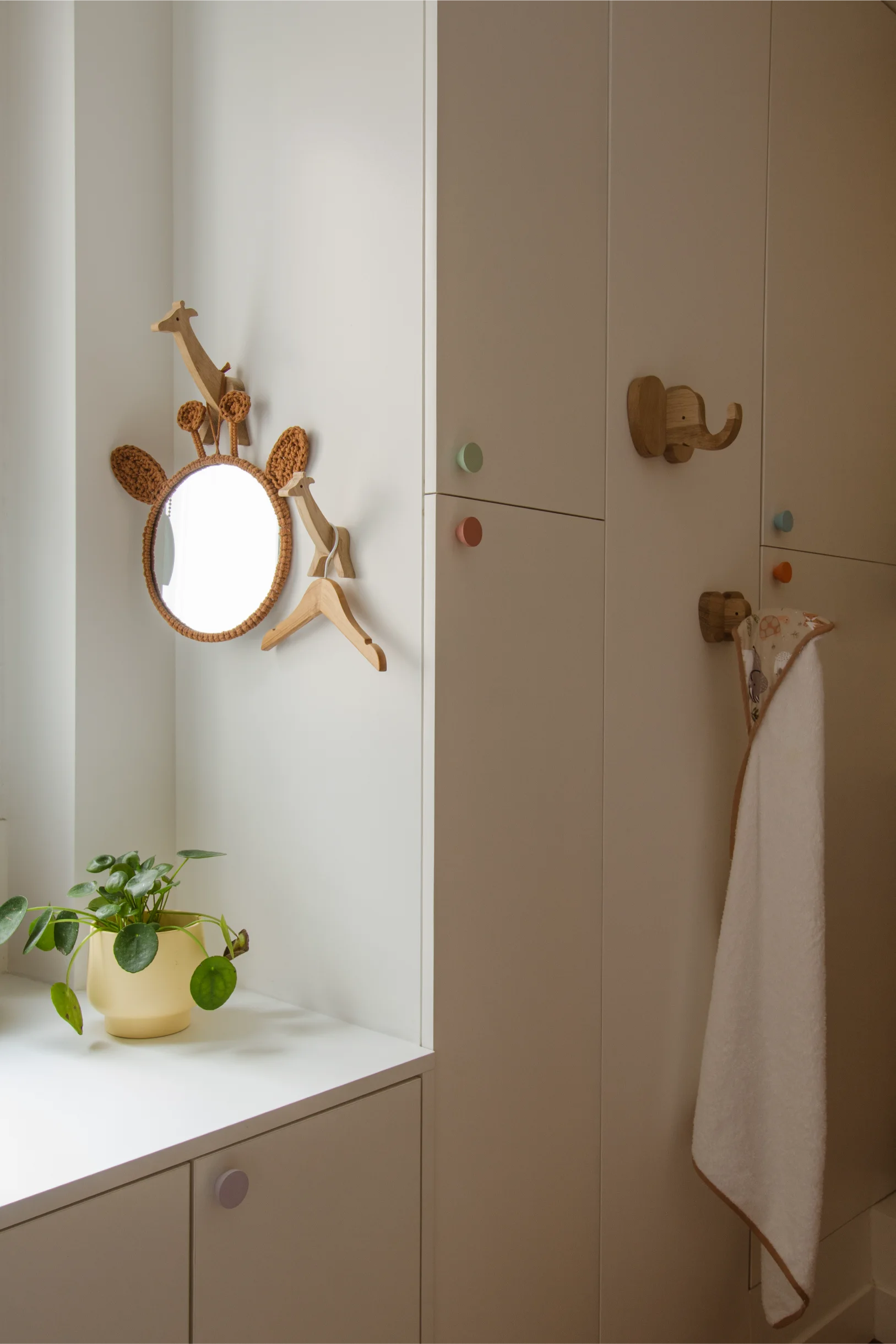 Coin chambre d'enfant beige clair avec un miroir rond sur le thème des girafes, un porte-manteau en bois en forme de girafe, une plante verte en pot et une serviette suspendue à des crochets en bois en forme d'éléphant et d'ours.