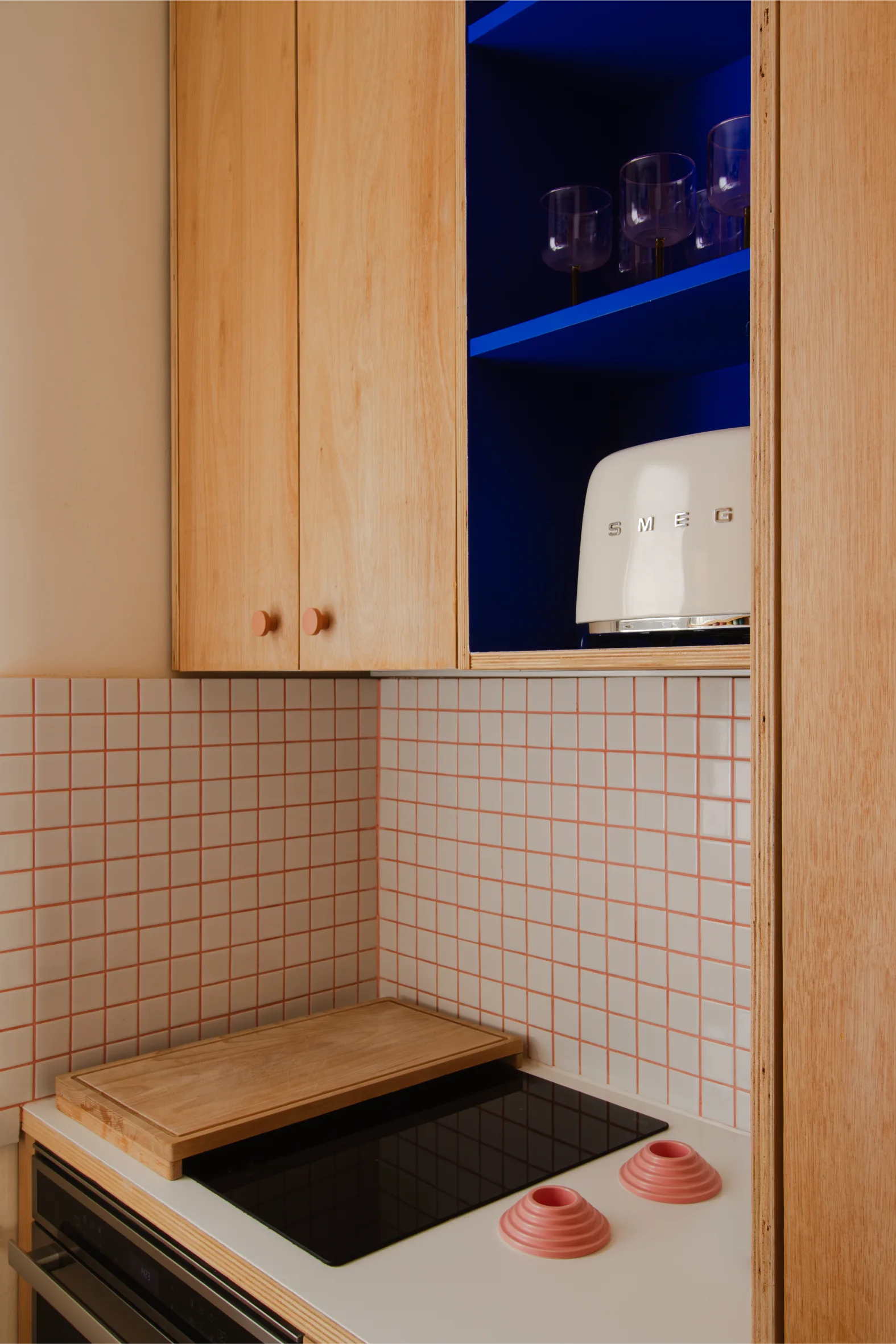 Coin cuisine moderne avec des armoires en bois, un dosseret en carreaux blancs avec du coulis rose, une plaque de cuisson électrique noire et un grille-pain SMEG blanc sur une étagère bleue.