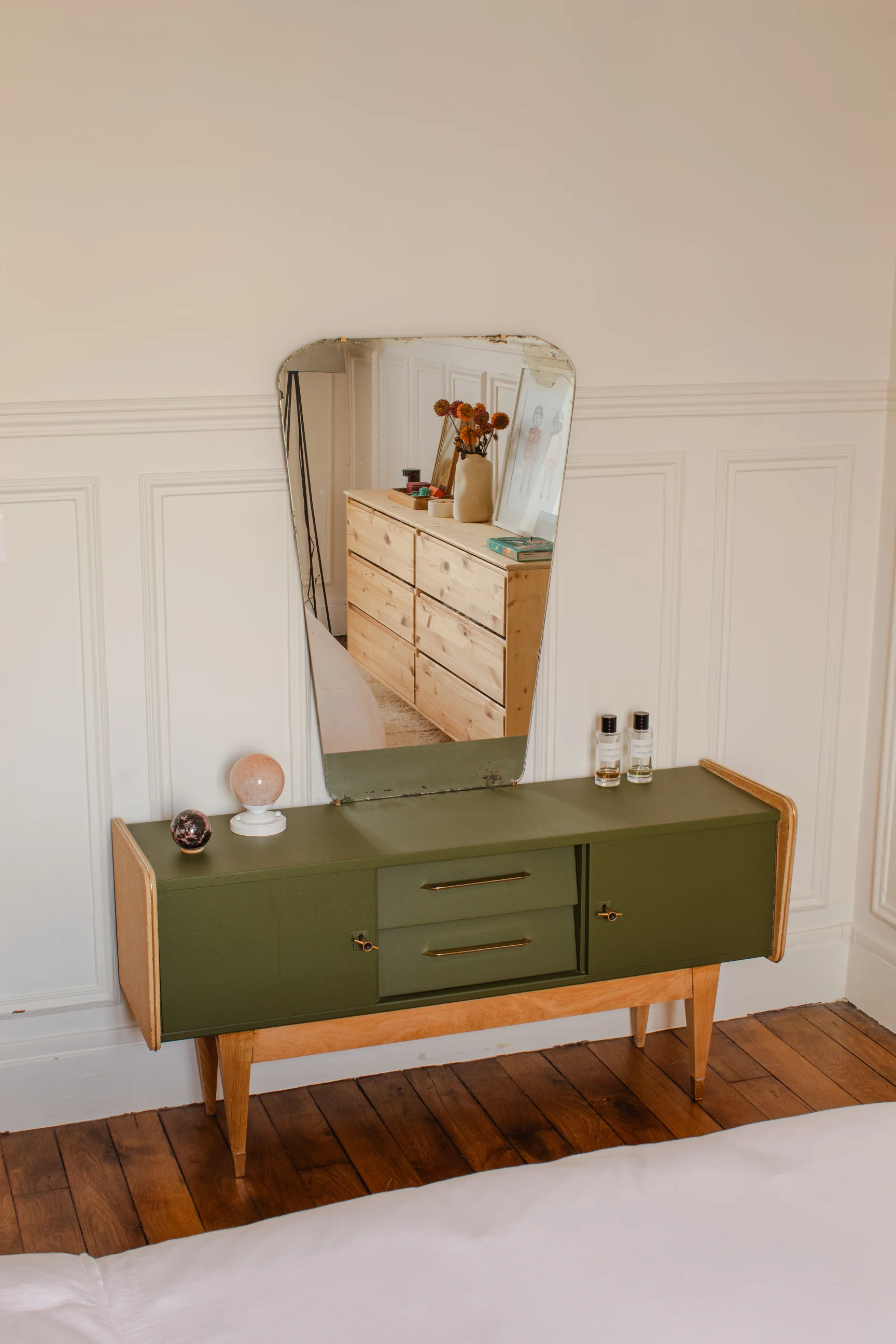 Commode moderne verte du milieu du siècle avec pieds en bois, surmontée d'un miroir vintage, de sphères décoratives et de deux petites bouteilles dans une pièce faiblement éclairée avec un sol en bois.