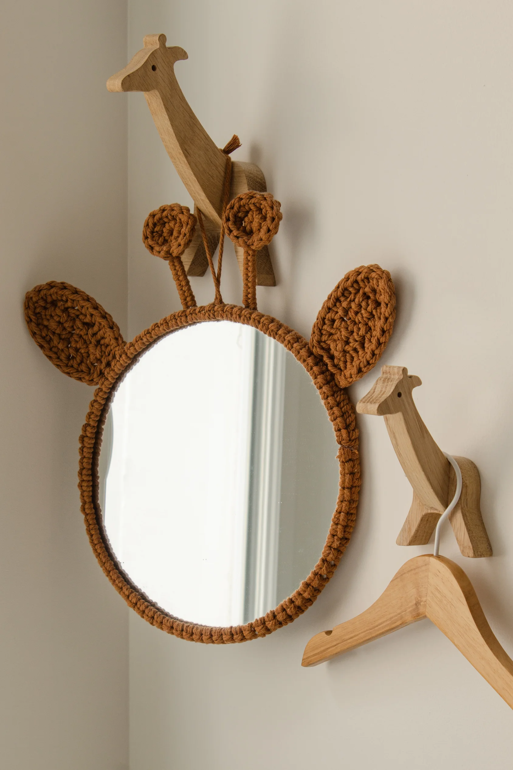 Miroir rond encadré d'un tissu crocheté marron conçu pour ressembler à des oreilles et des cornes de girafe, suspendu à côté de deux crochets muraux en bois en forme de girafe avec un cintre en bois.