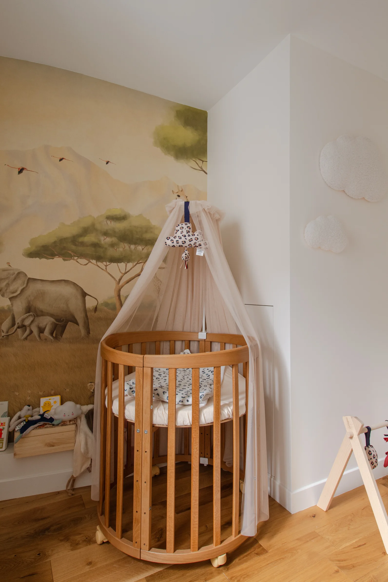 Lit bébé rond en bois avec baldaquin blanc transparent dans une chambre d'enfant décorée d'une fresque murale représentant des éléphants et des arbres.
