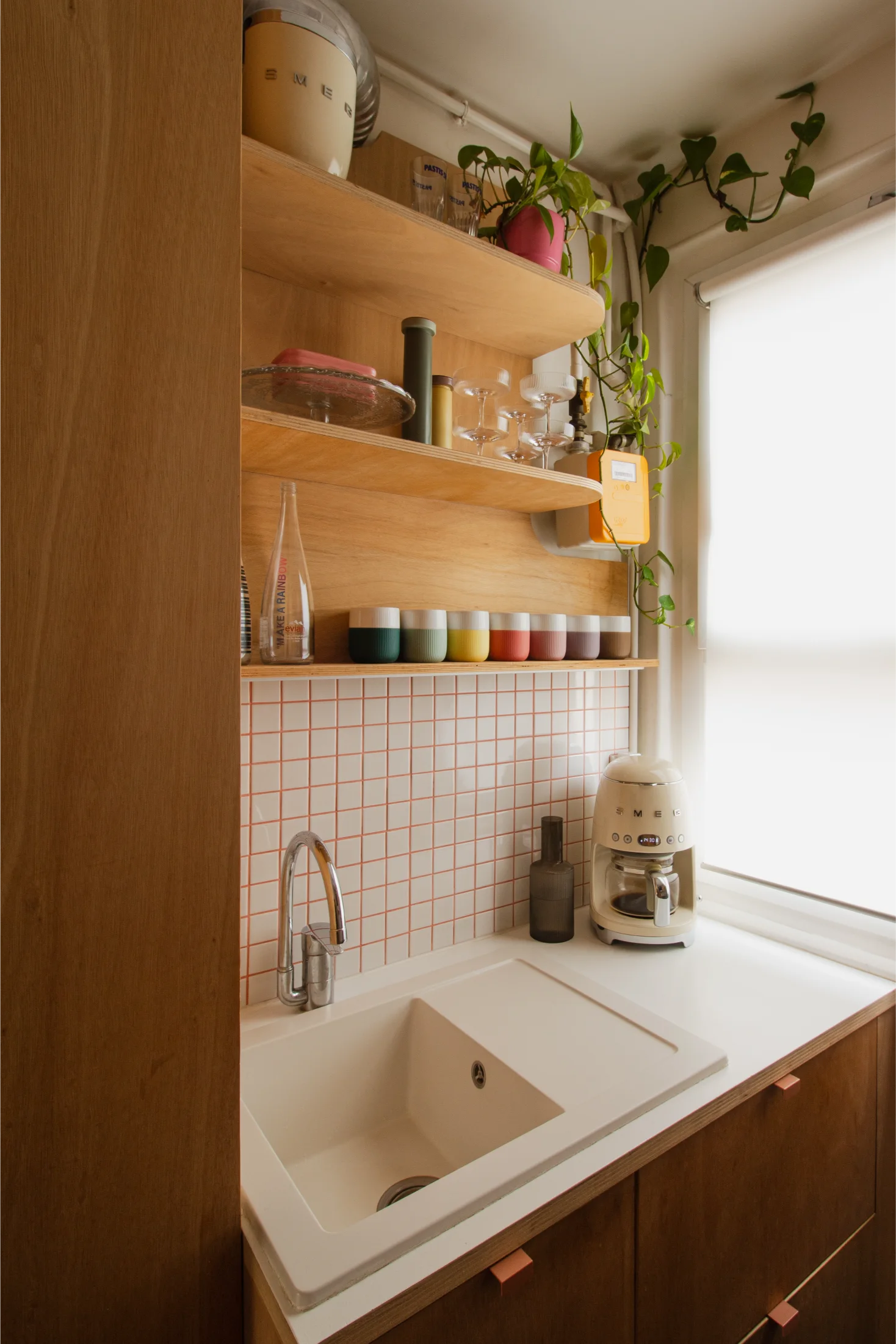 Coin cuisine moderne avec évier blanc, tasses en céramique colorées sur des étagères en bois, cafetière SMEG couleur crème et plante retombante près d'une fenêtre équipée de stores blancs.