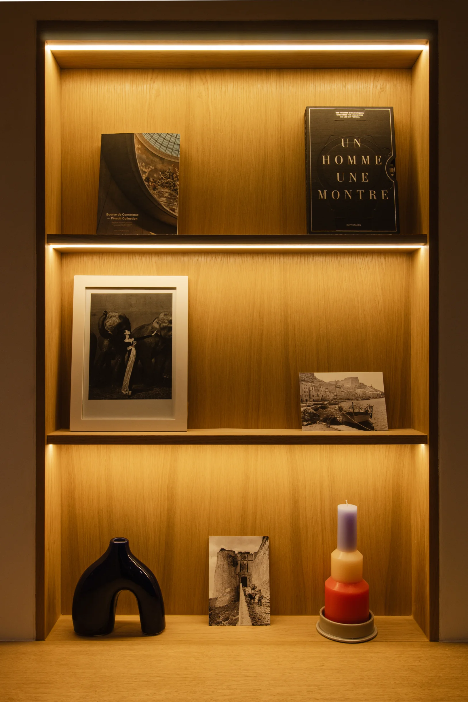 Étagère en bois avec éclairage chaleureux, sur laquelle sont disposés des livres, des photos noir et blanc encadrées, un vase en céramique bleu et une bougie multicolore à plusieurs couches.