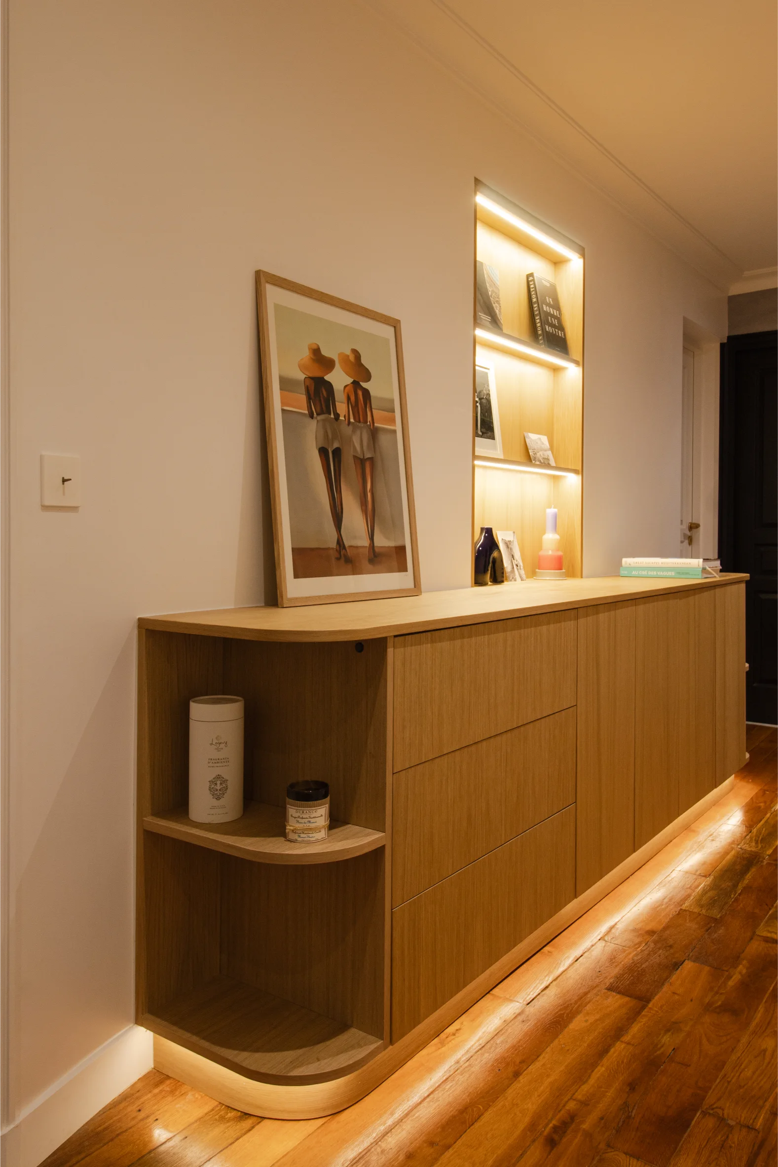 Buffet en bois avec coin arrondi, cadre accroché dessus, objets décoratifs sur l'étagère ouverte et étagères encastrées éclairées au-dessus.