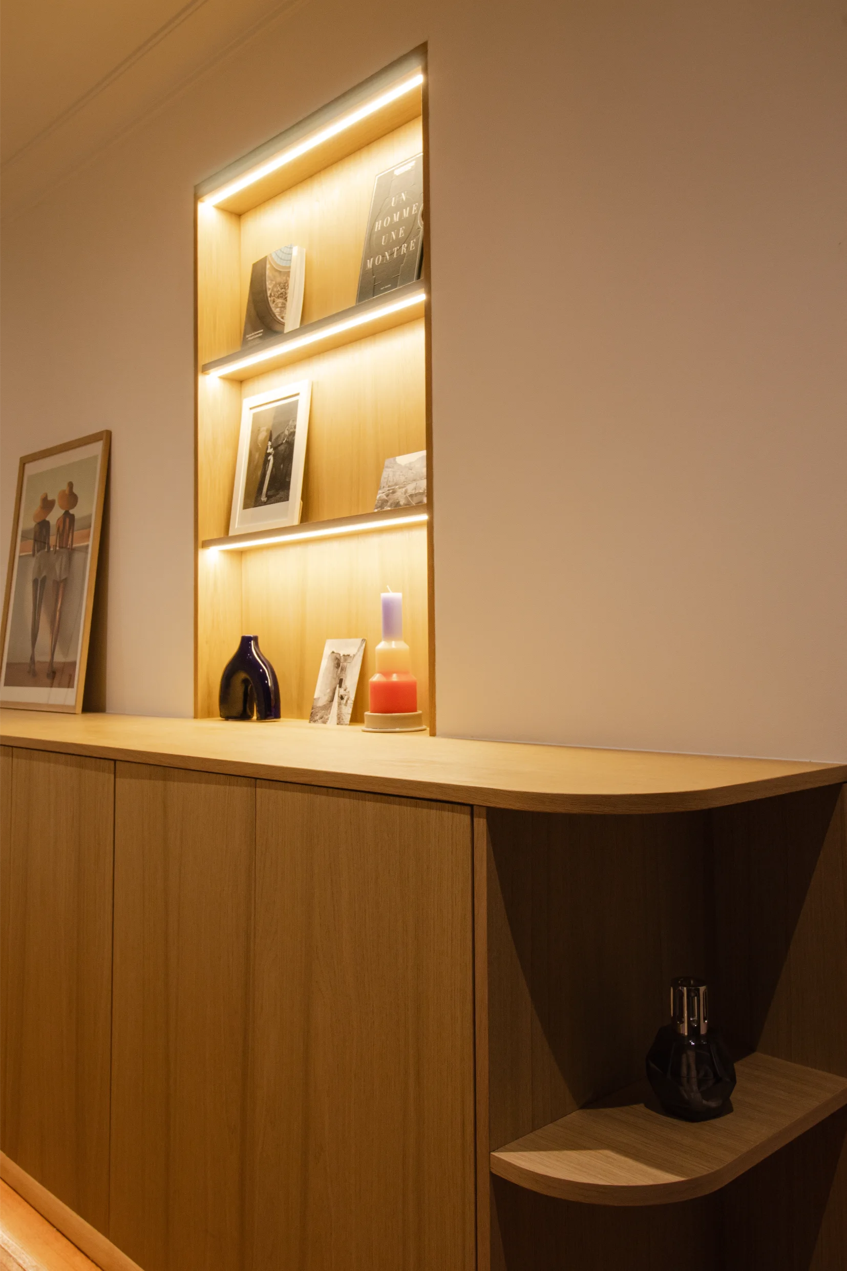 Armoire en bois avec étagères éclairées intégrées exposant des livres, des photos encadrées, un vase en céramique bleu-violet et une bougie multicolore.