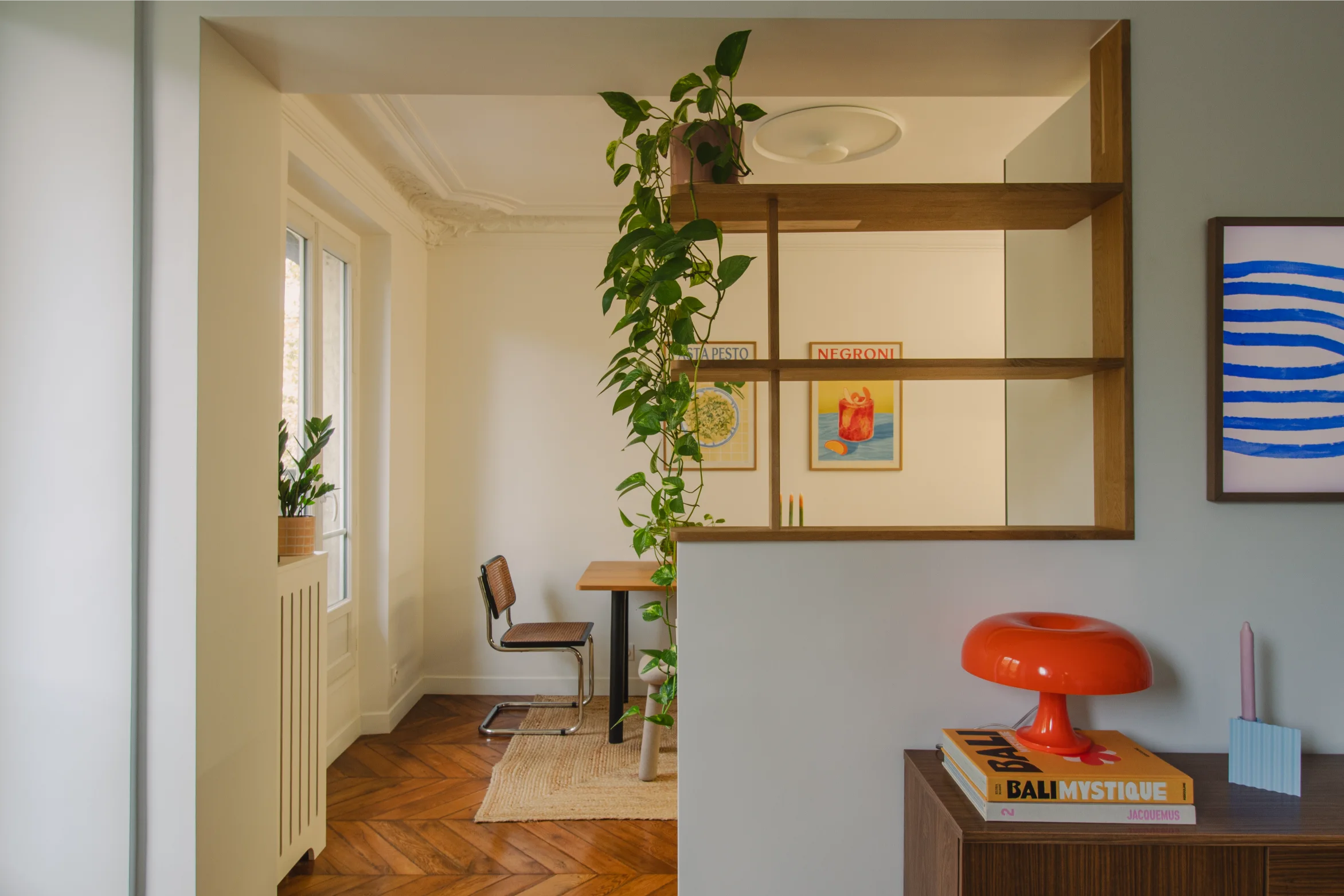 Intérieur moderne avec parquet en bois, plante verte suspendue, petite table à manger avec chaise, affiches décoratives et lampe rouge en forme de champignon posée sur un meuble.