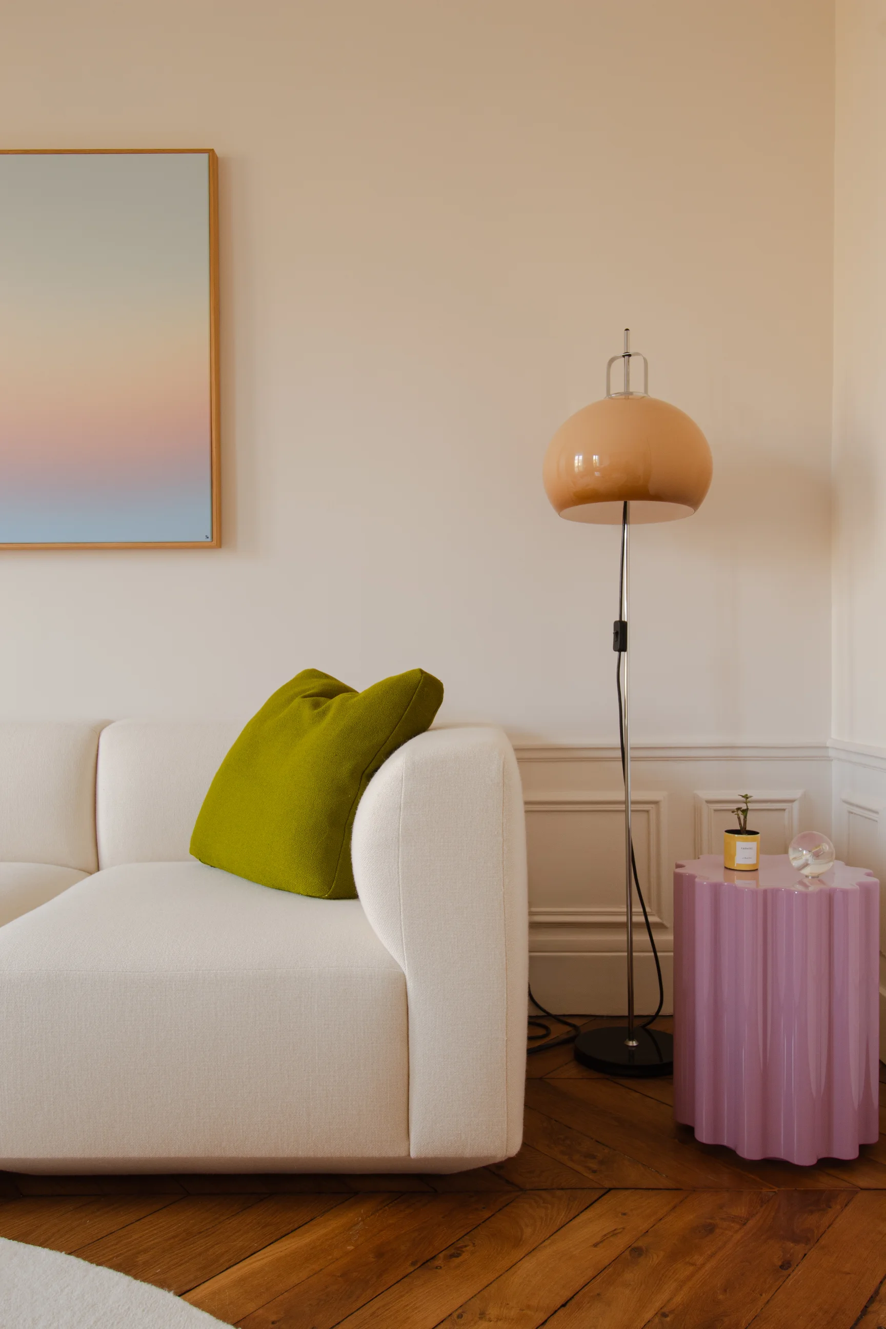 Canapé beige moderne avec un coussin vert à côté d'une table d'appoint lavande et d'un lampadaire pêche dans un salon avec parquet.