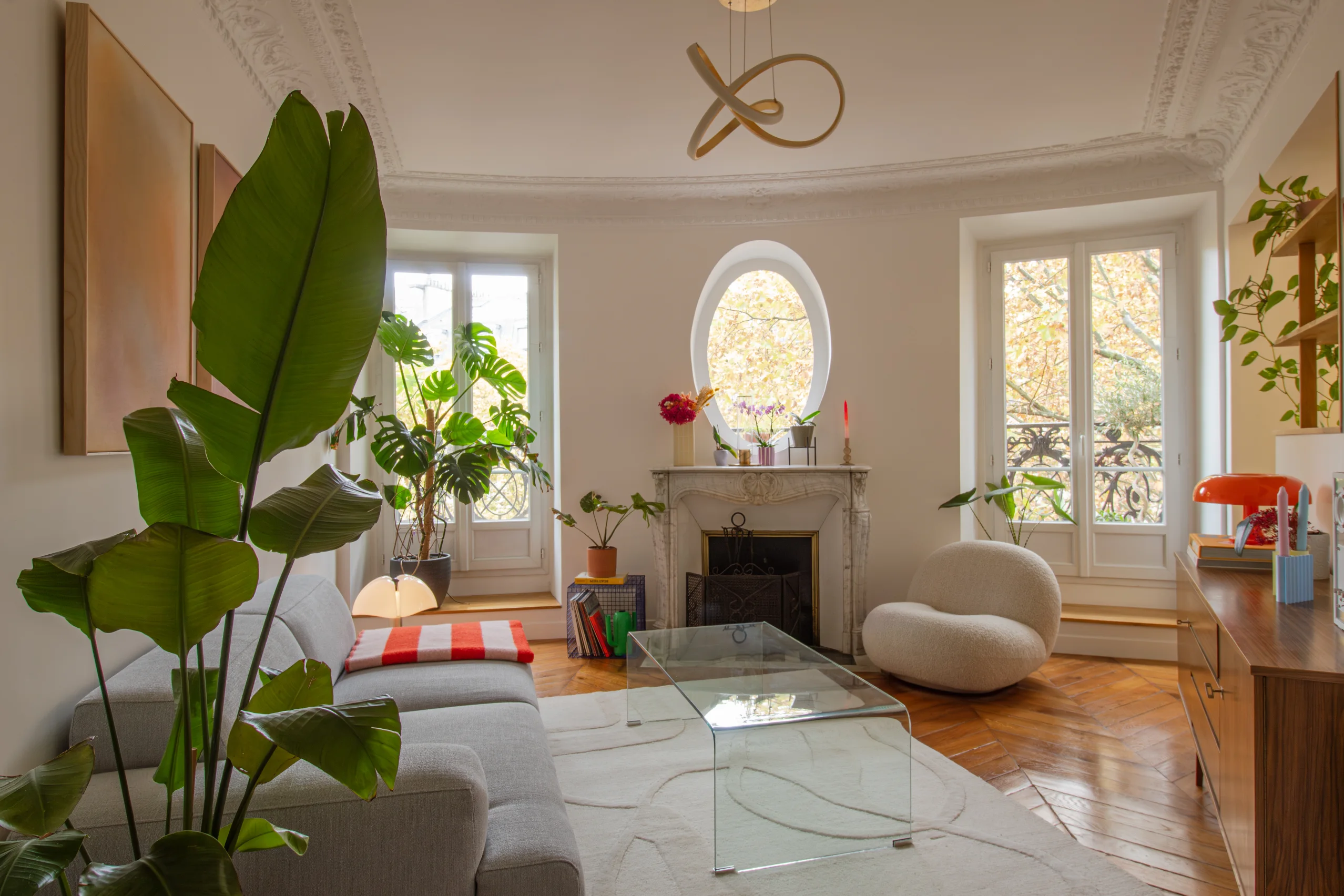 Salon moderne avec un canapé gris, un fauteuil blanc texturé, une table basse en verre, de grandes plantes vertes et une cheminée décorative sous une fenêtre ovale.