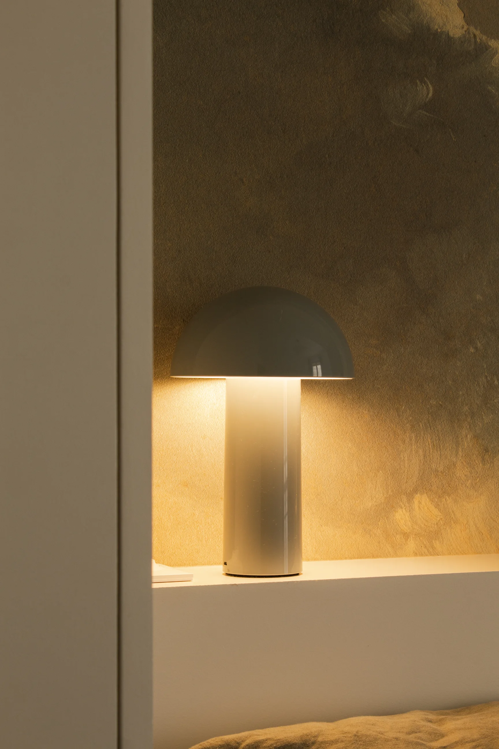 Lampe de chevet moderne avec un abat-jour bombé diffusant une lumière chaude sur une étagère blanche contre un mur beige texturé.